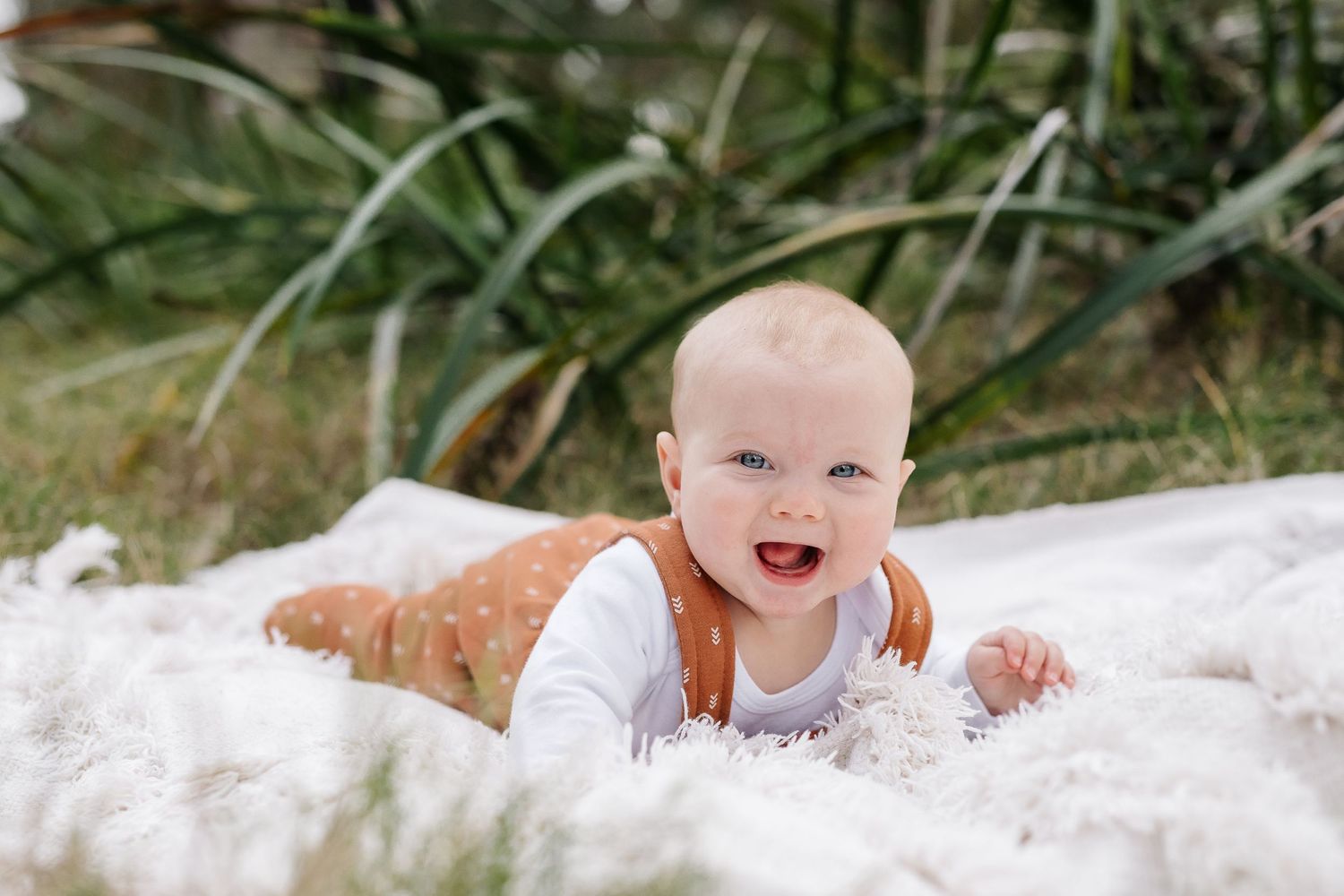 Smiling baby in orange overalls lying on white blanket outdoors.