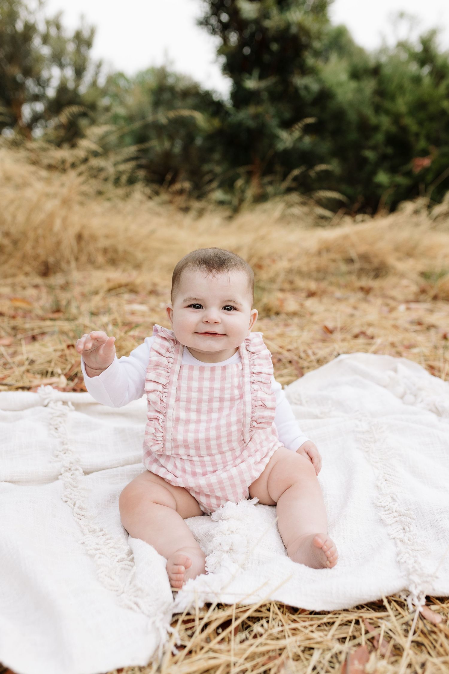 Baby in pink gingham romper sits on white blanket outdoors surrounded by dry grass.