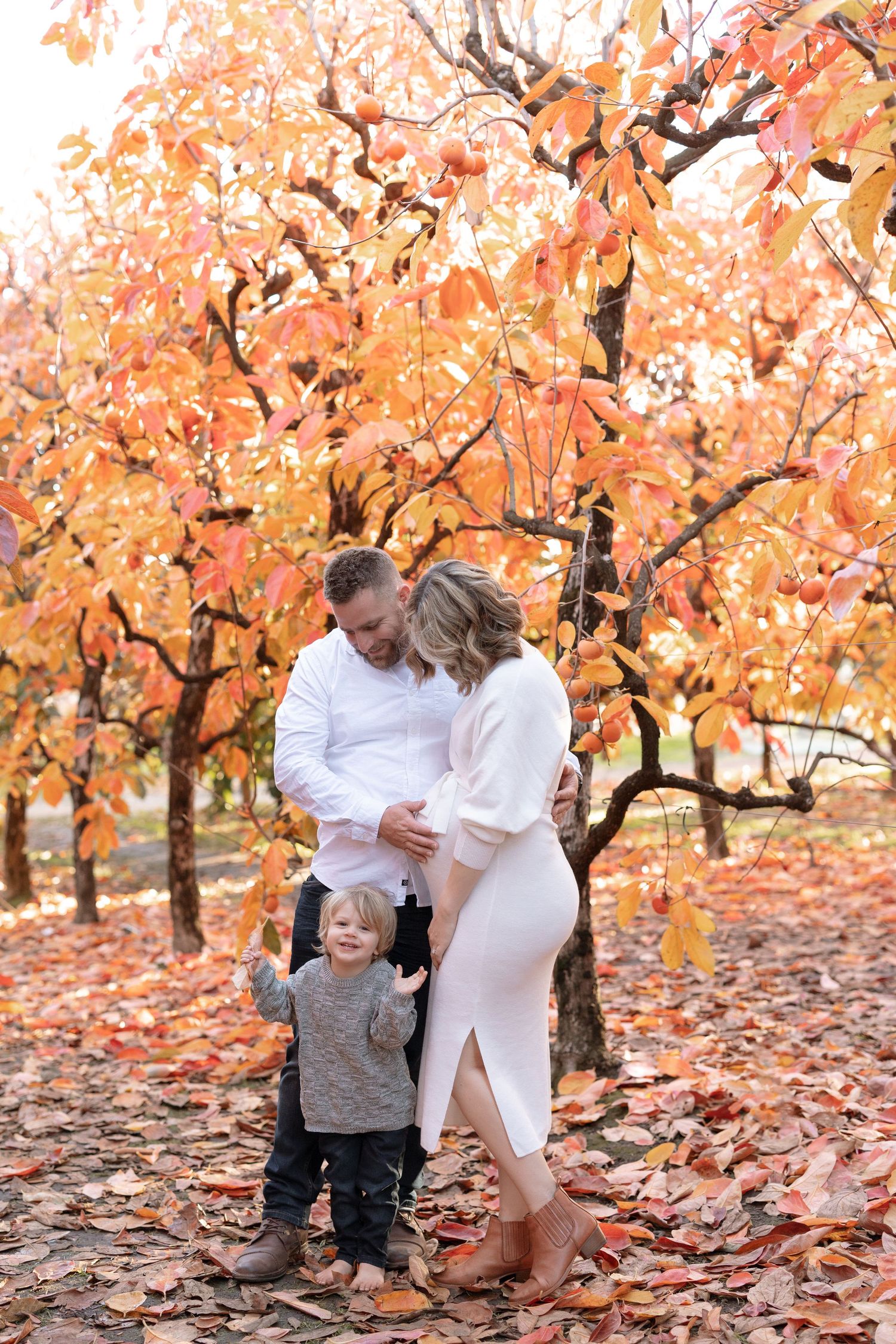 Family portrait under orange autumn trees with fallen leaves on the ground.
