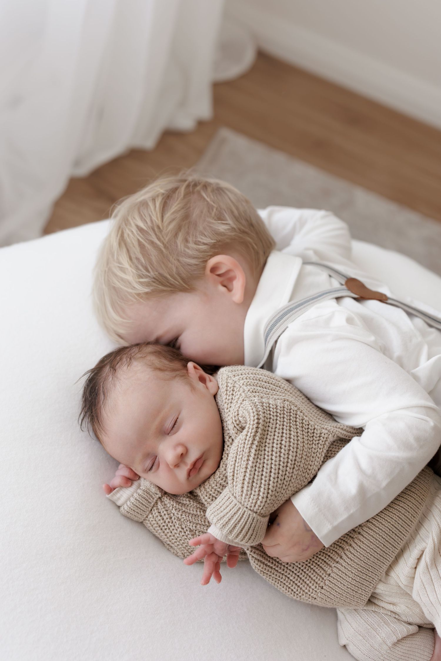 Heartwarming moment as siblings cuddle together on white bedding in natural light.