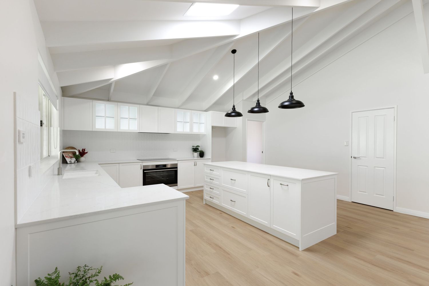 Modern white kitchen interior with vaulted ceiling, pendant lights, hardwood floors and minimalist cabinetry design.