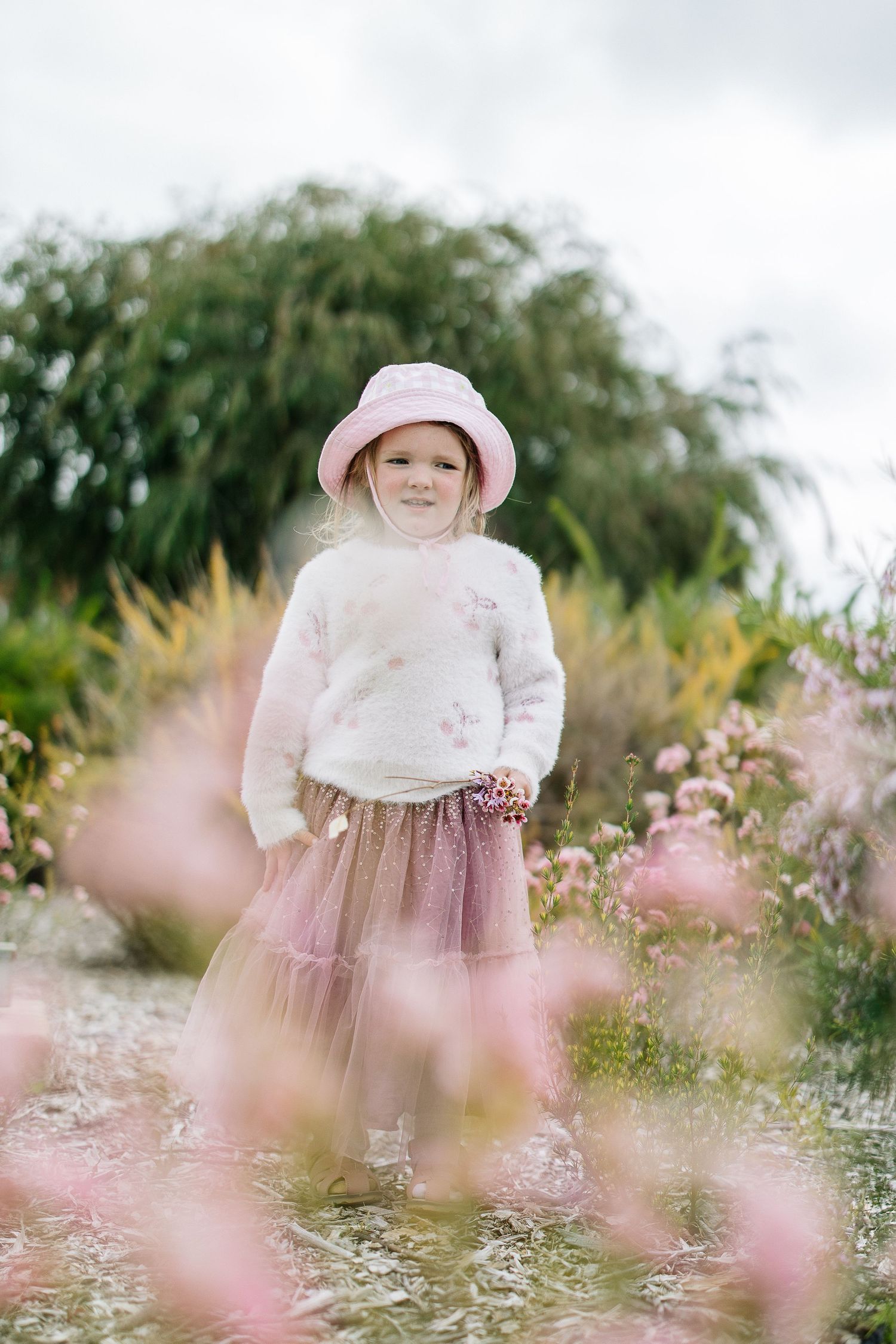 Garden portrait with pink flowers and matching hat against willow trees.
