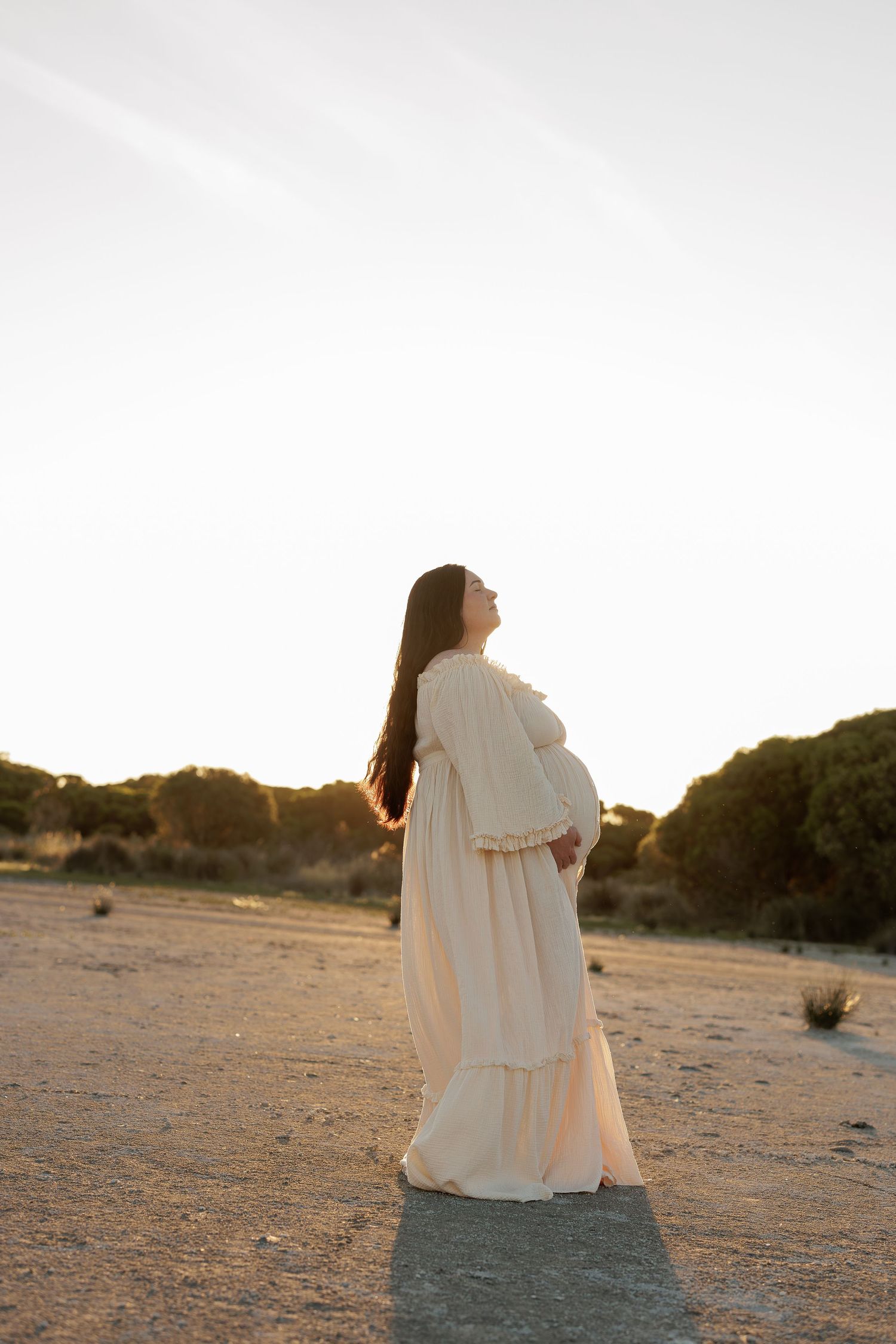 A pregnant woman in a flowing white dress stands in a desert landscape at sunset casting a long shadow.