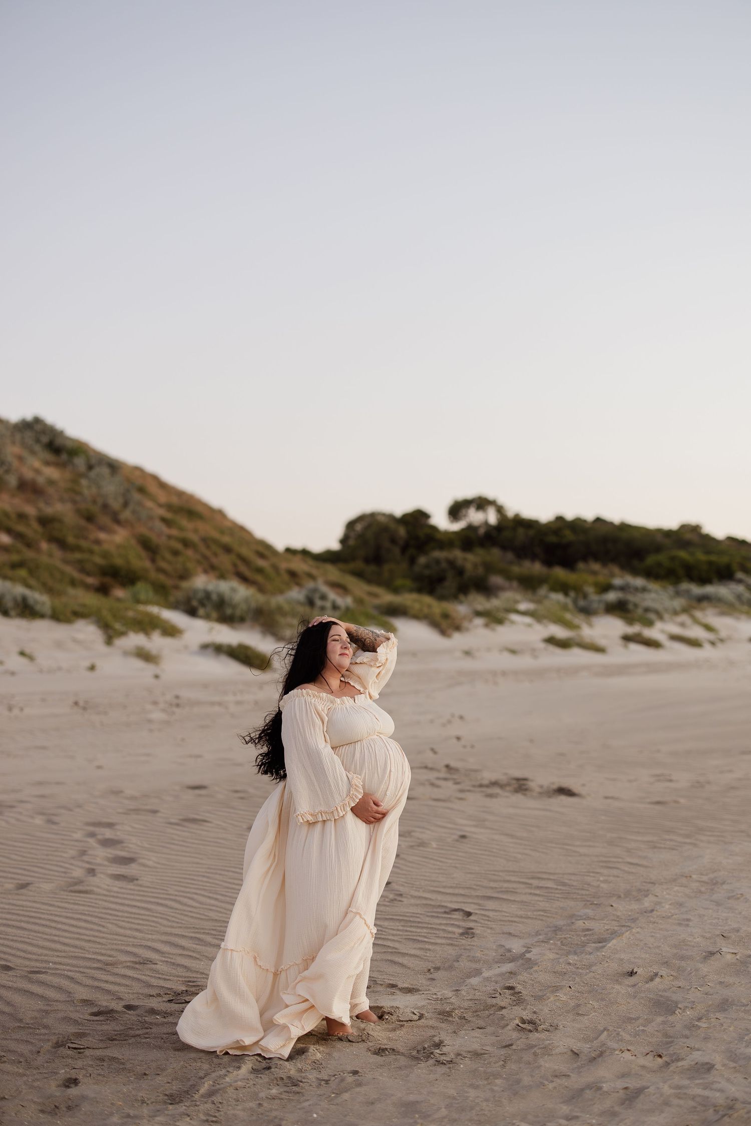 Woman in flowing white dress poses gracefully on sandy beach at sunset.