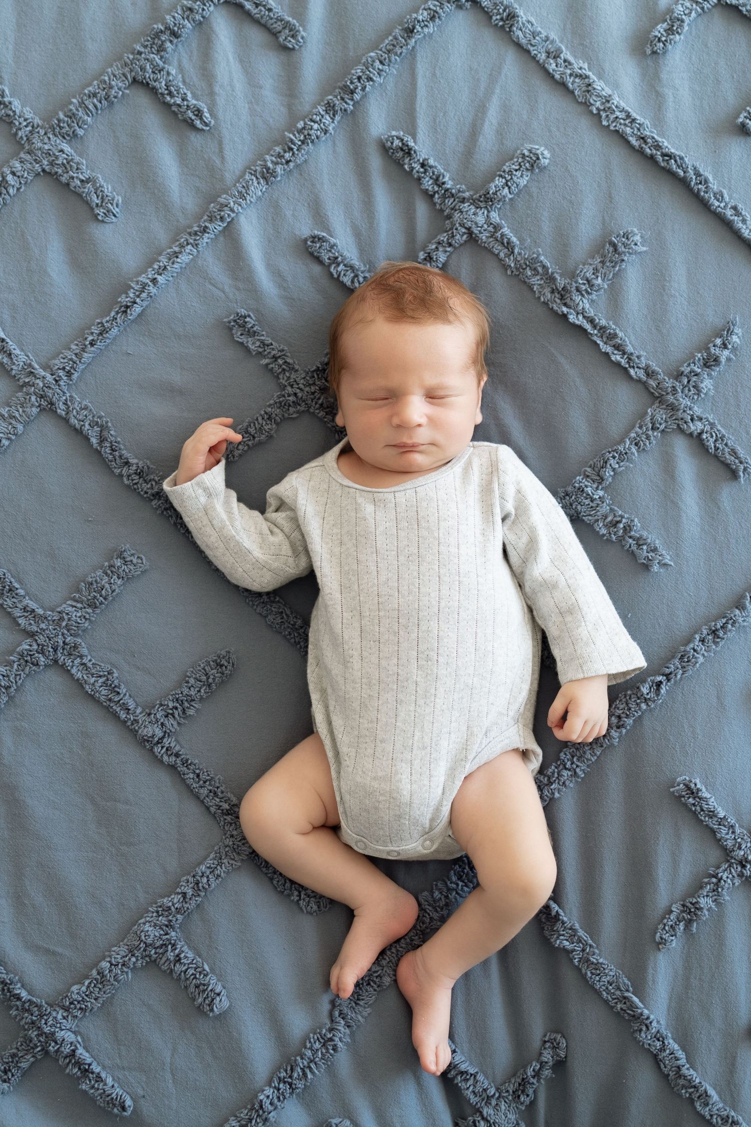 Sleeping newborn in white onesie lying on gray textured bedding with diamond pattern.