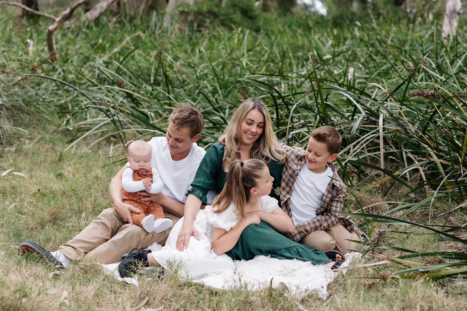 Candid family photo of five people sitting together on picnic blanket in tall grass.