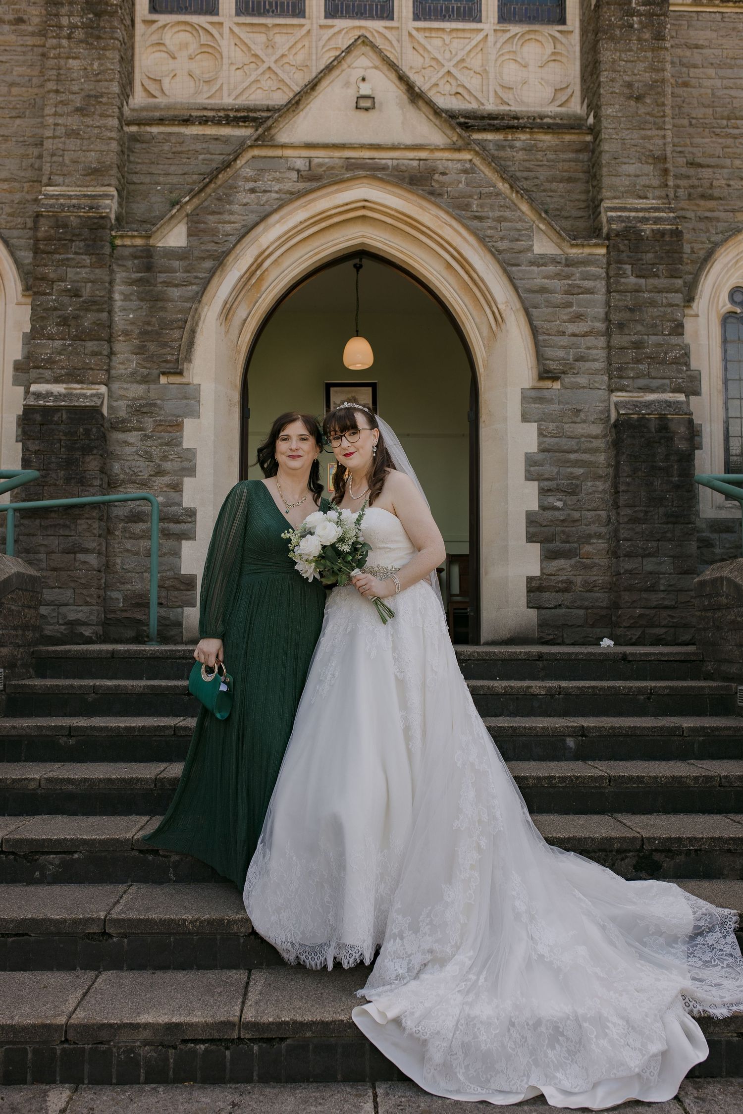 Bride in white gown and woman in green dress pose on church steps.