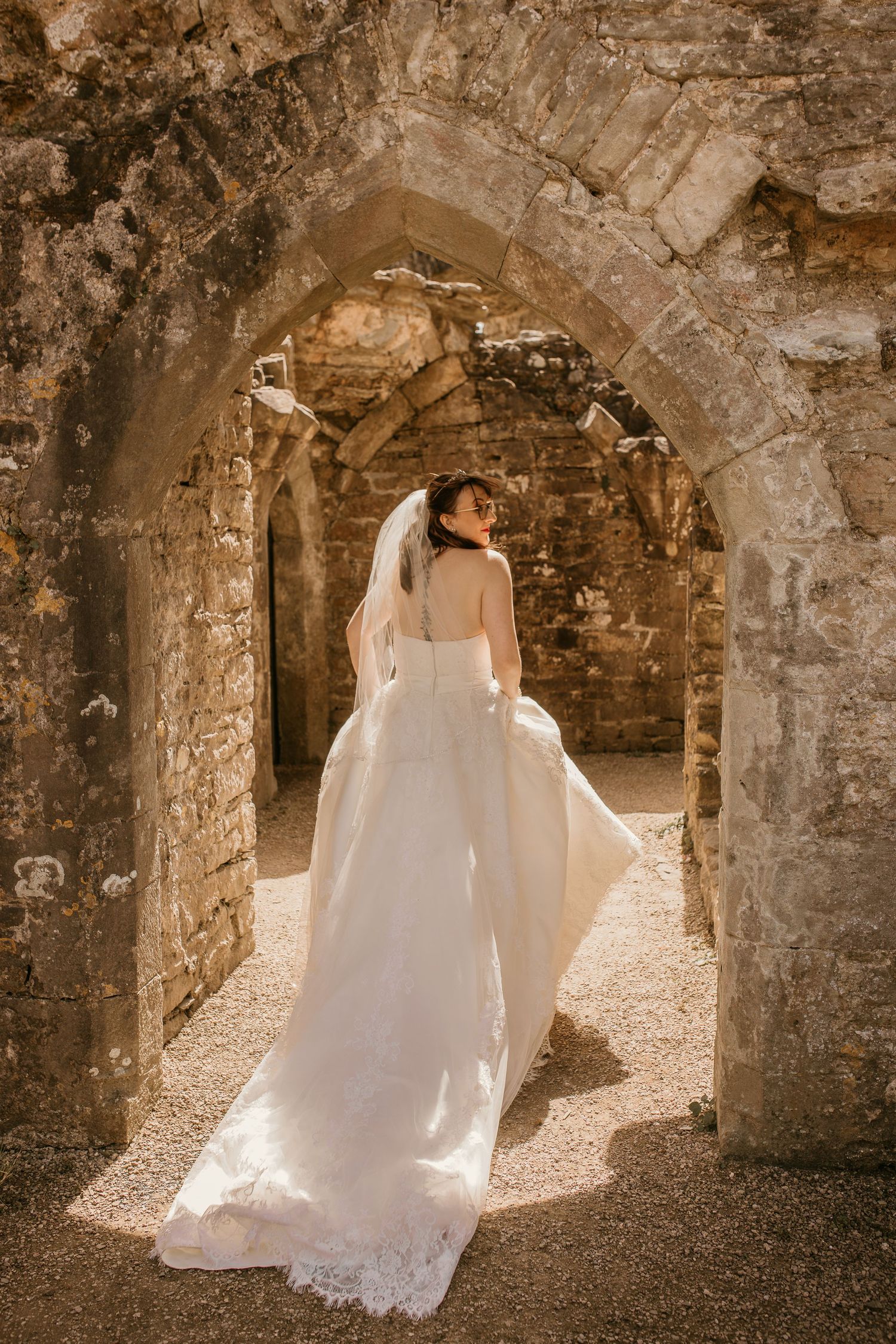 Bride in white gown and veil stands in stone archway of ancient ruins, viewed from behind.