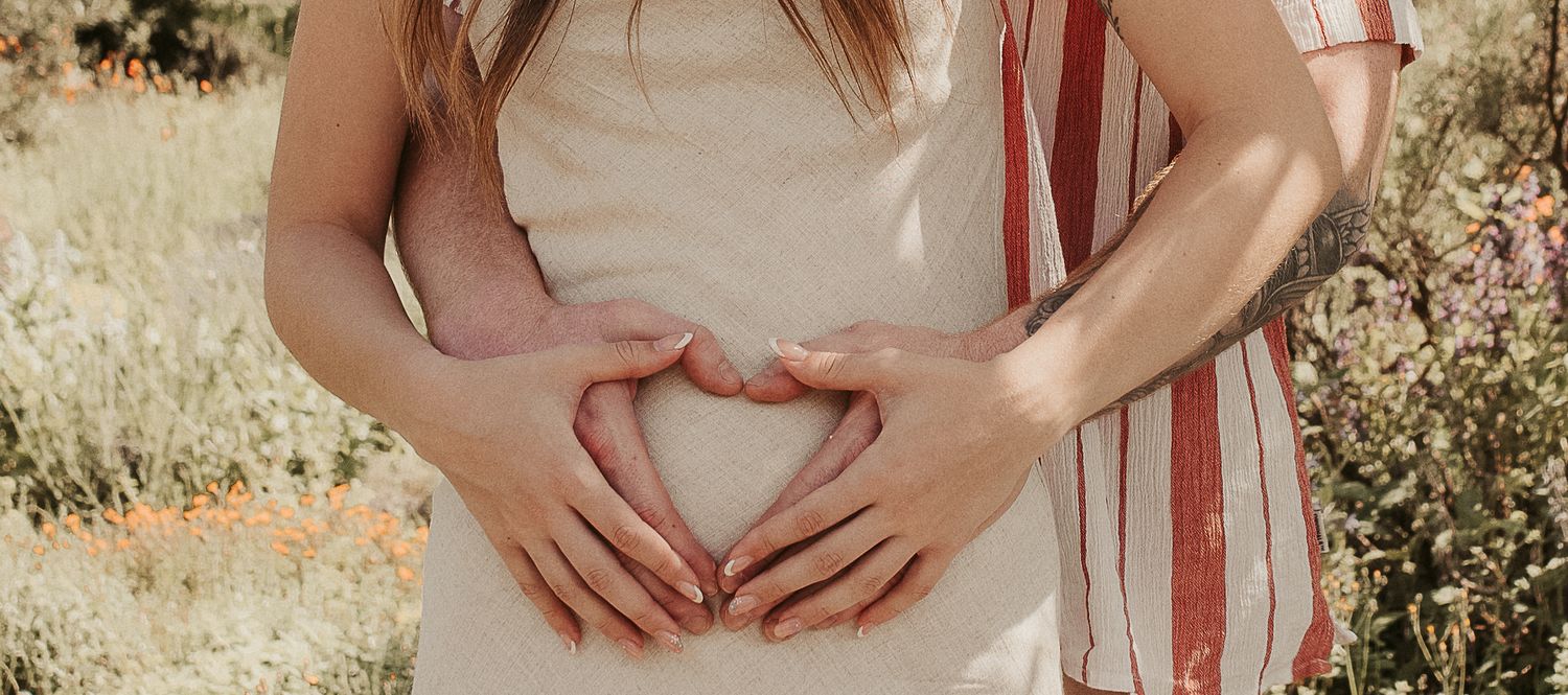 Couple forming a heart shape with their hands over a pregnant belly outdoors in a sunny field.