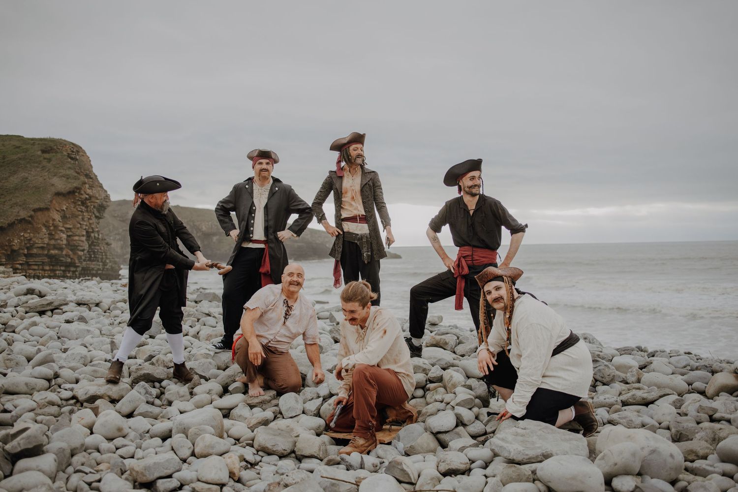 People in vintage costumes climbing over rocky coastal terrain on a cloudy day.