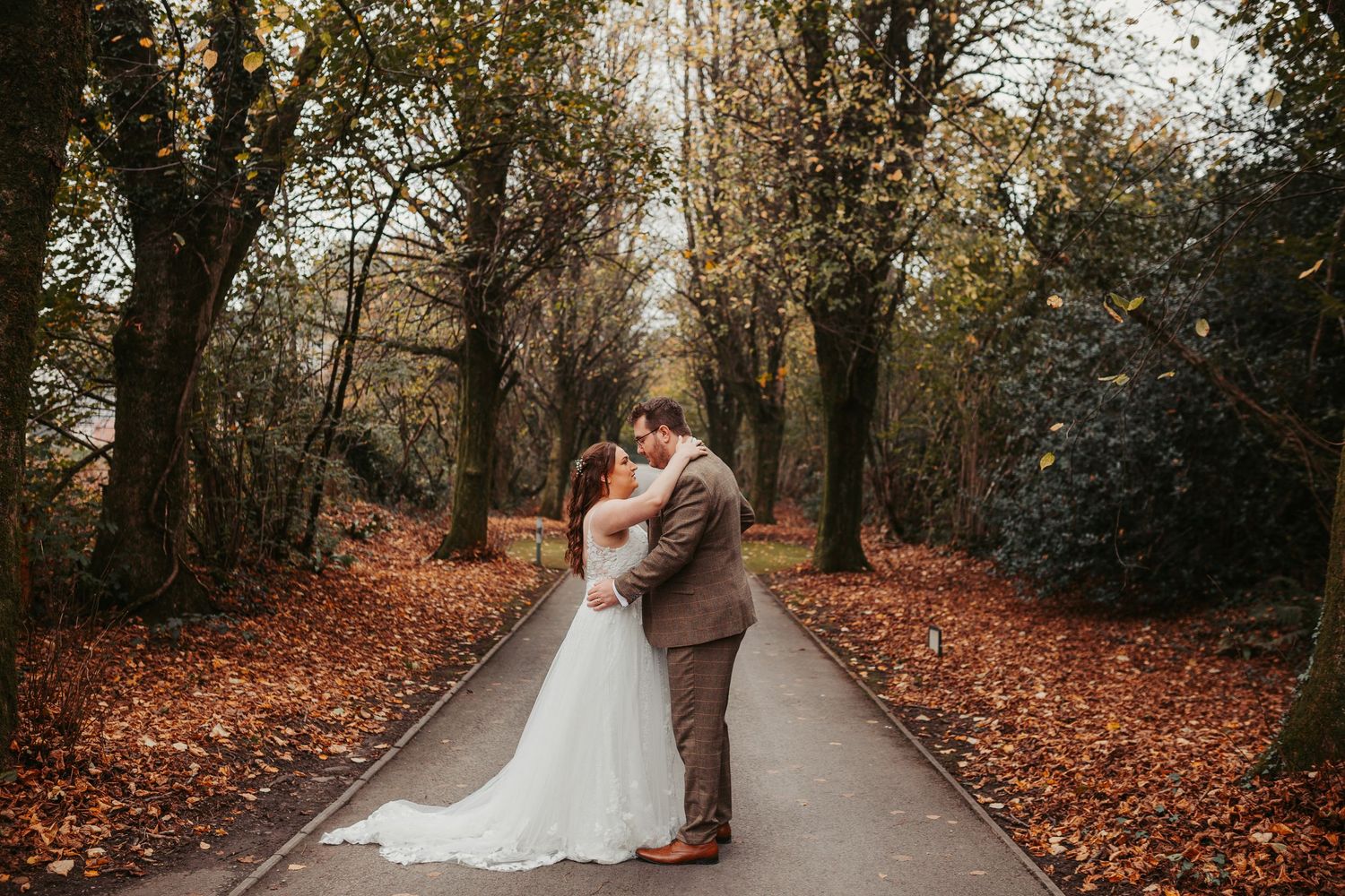 A romantic autumn wedding photoshoot on a tree-lined path covered in fallen orange leaves.