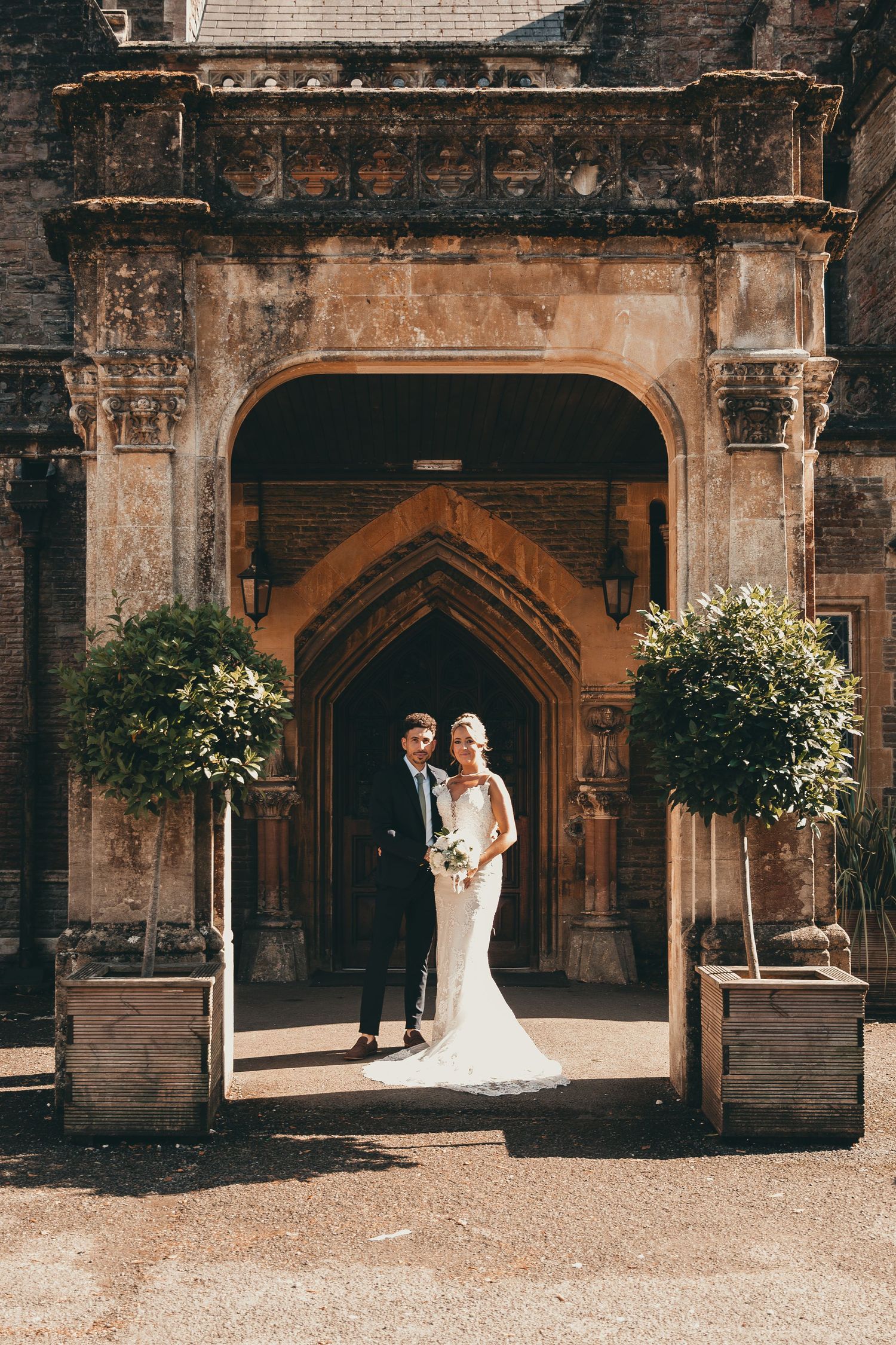 A wedding couple standing under an ornate stone archway with potted plants at a historic building entrance.