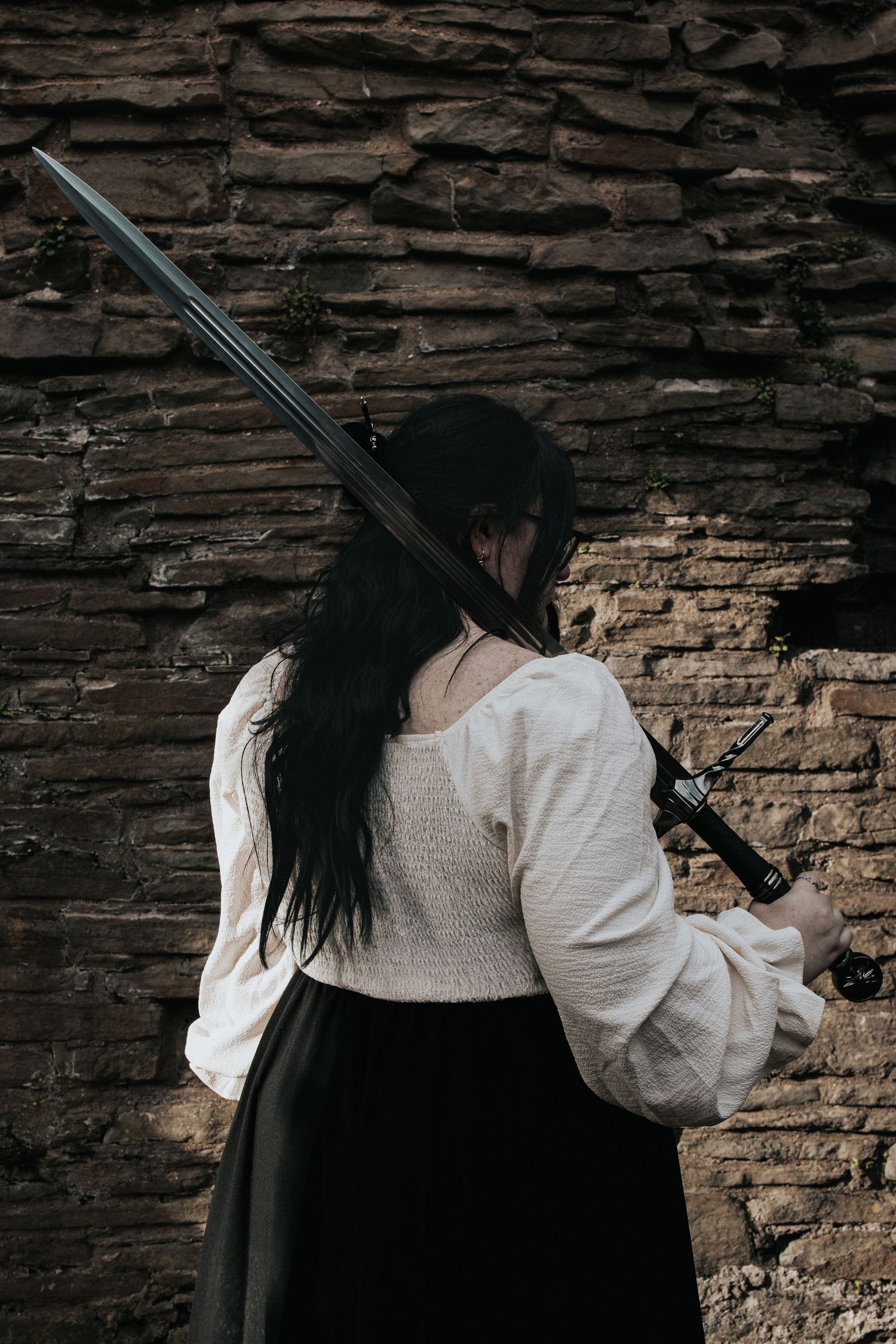 A dramatic side view of someone holding a sword against an ancient stone wall.