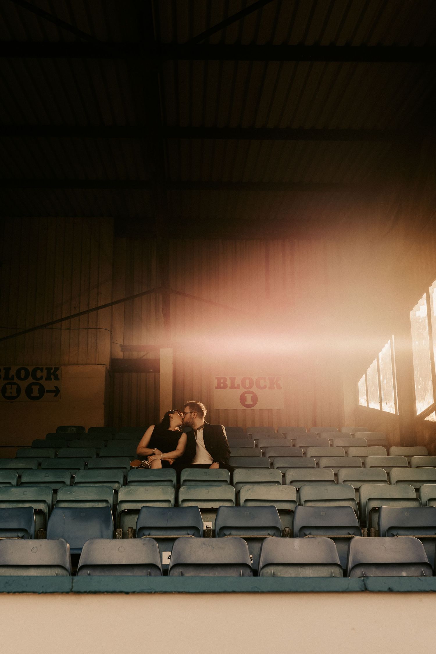 Couple shares a kiss in sunlit stadium seating area.