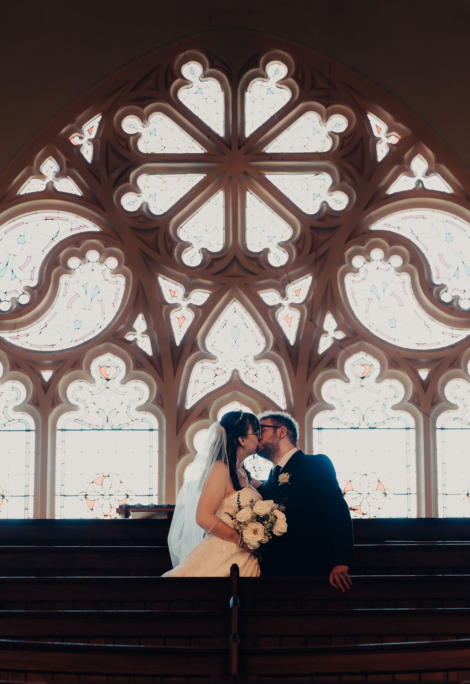 Bride and groom kissing in front of ornate Gothic rose window in church pew, bride holding white floral bouquet.