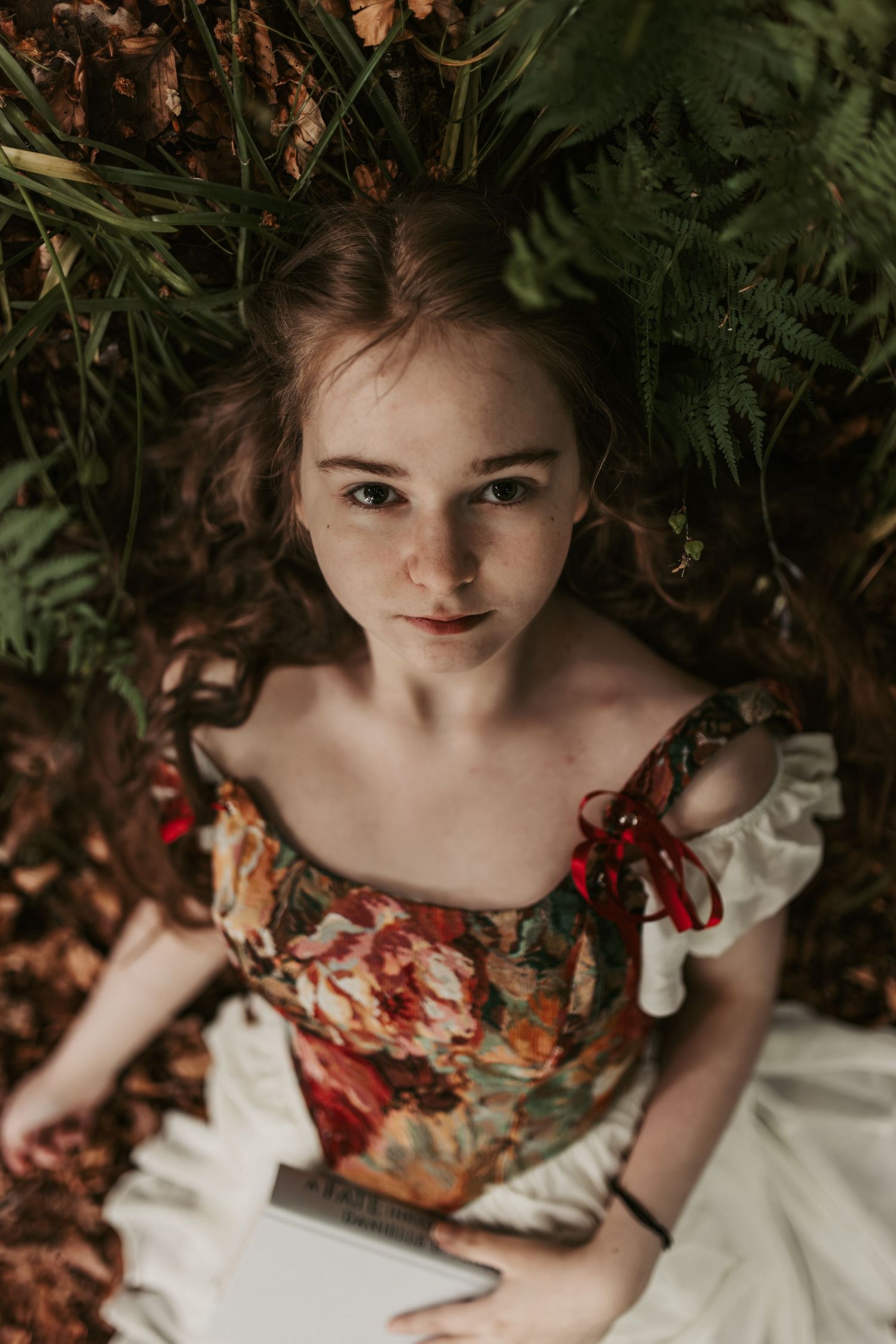 Young woman in floral off-shoulder dress lying among ferns and autumn leaves, gazing upward holding a book.