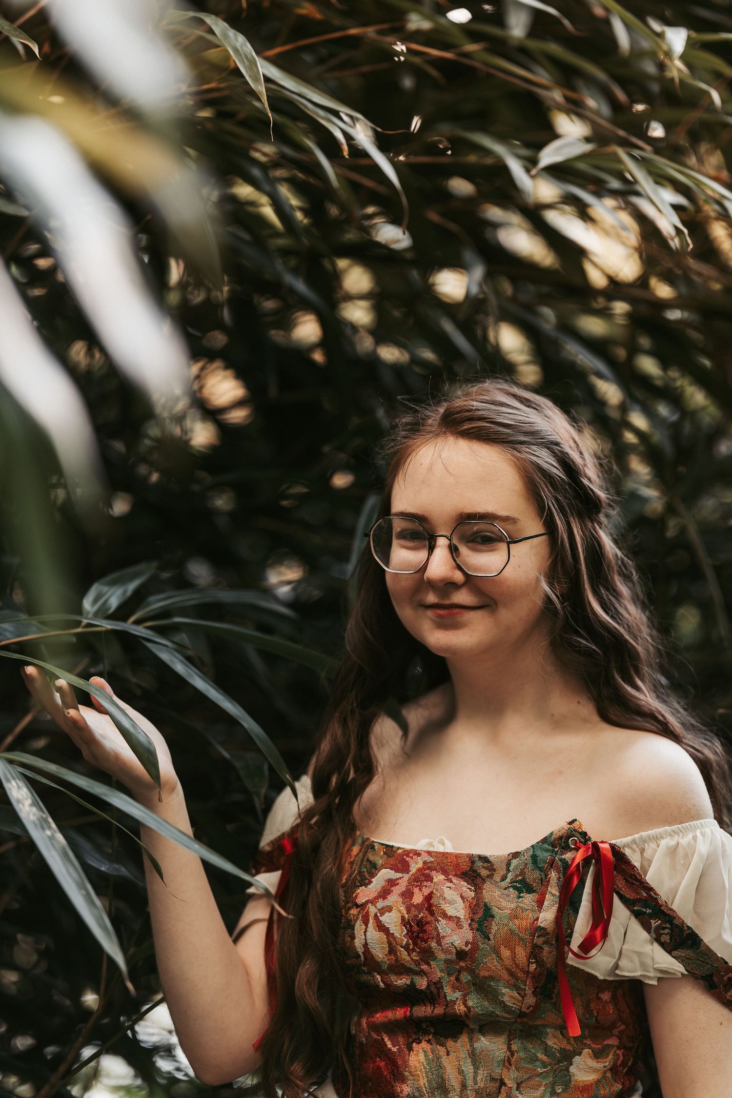 Young woman with glasses and long wavy hair wearing a floral off-shoulder dress, standing among lush green tropical plants.
