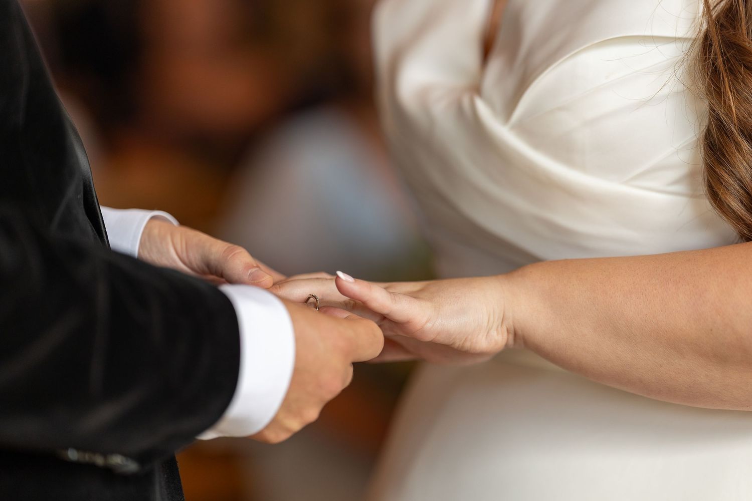 Close-up of hands exchanging wedding rings during a marriage ceremony with bride in white dress and groom in black suit.