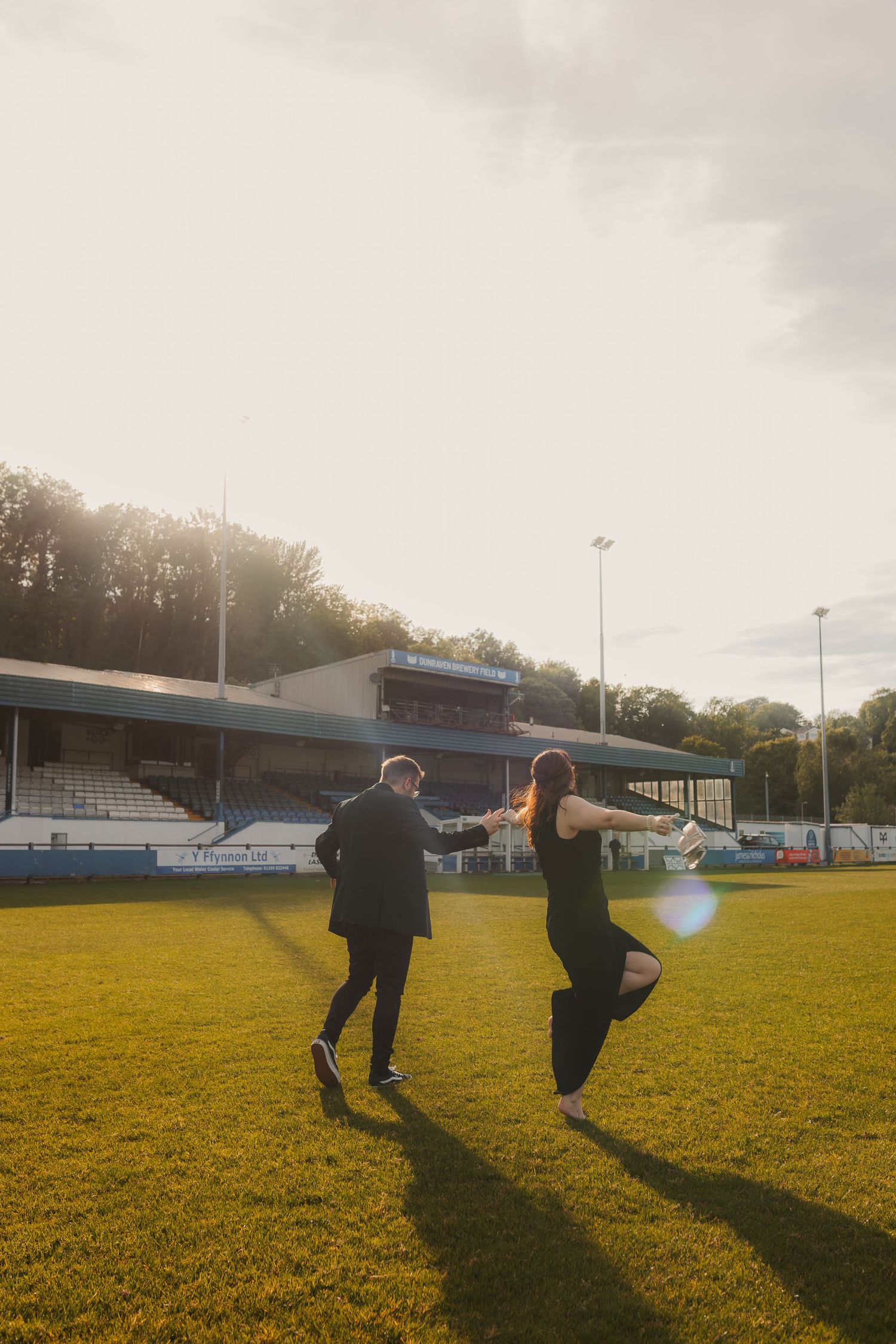 Couple runs hand-in-hand across a sunlit green football pitch.