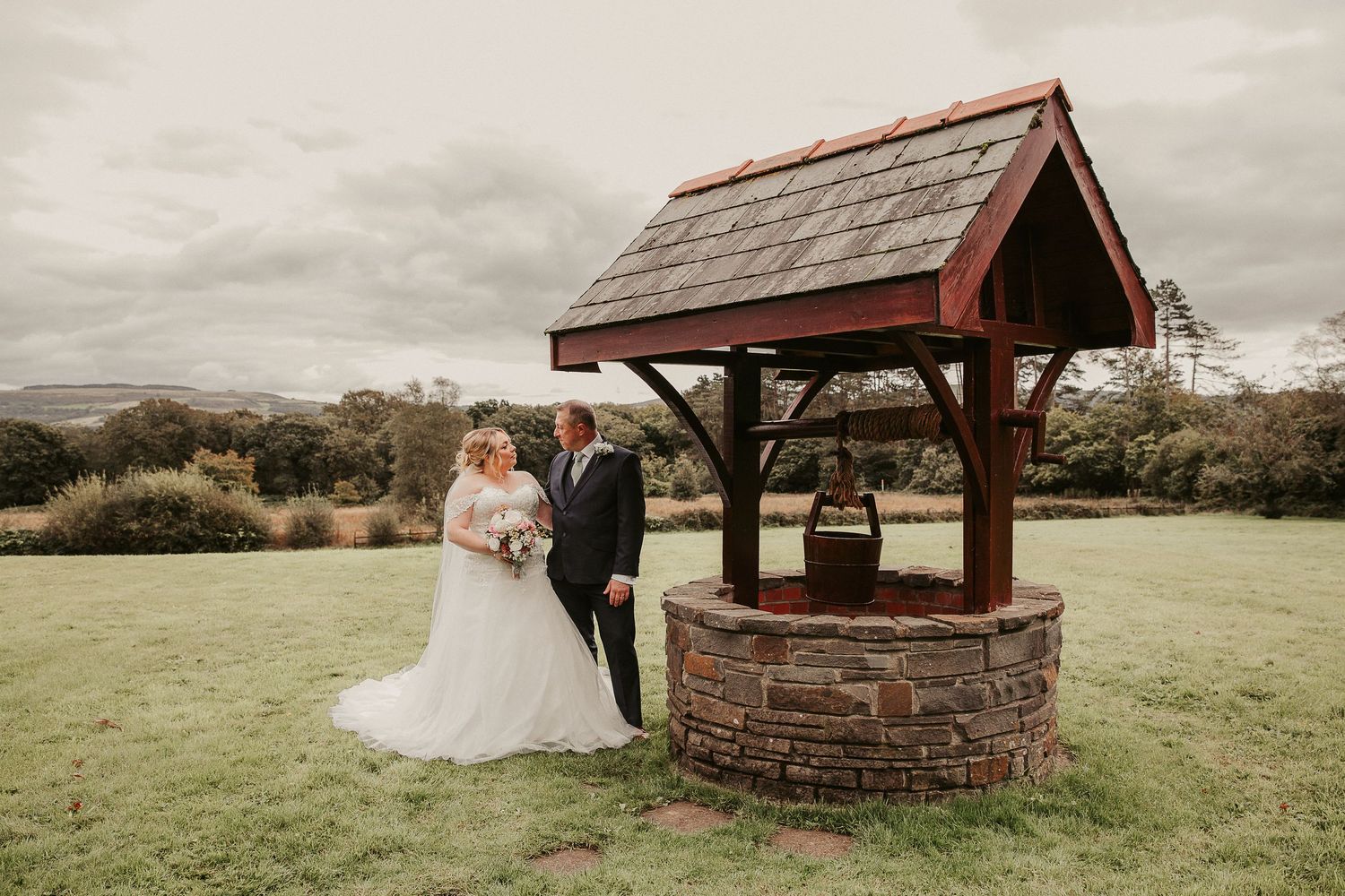Wedding couple poses near rustic stone well houses in a serene meadow landscape.