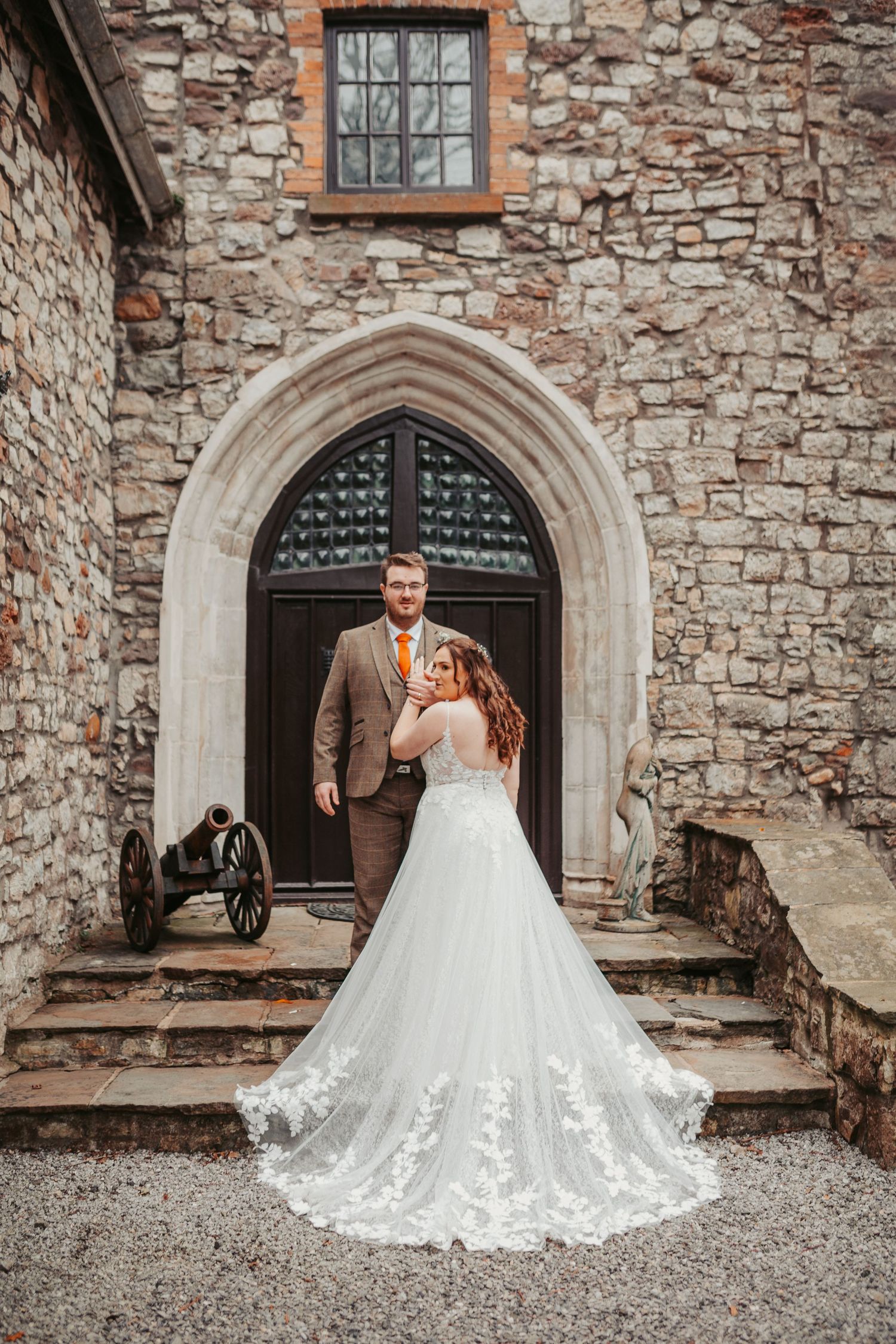 A wedding couple stands in front of a gothic stone archway with detailed lace train on display.