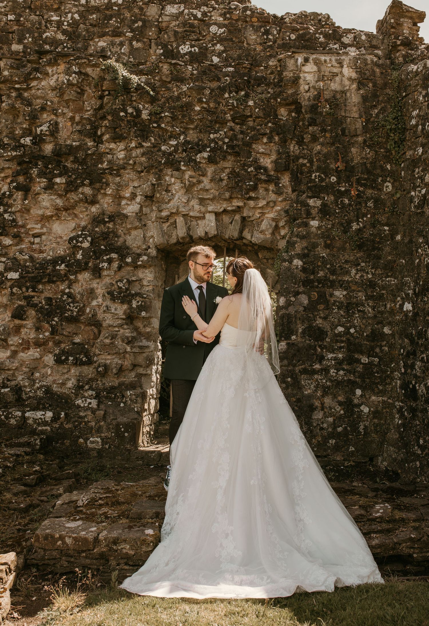 Bride and groom share a romantic moment in front of ancient stone ruins, bride wearing a flowing white lace gown with veil.