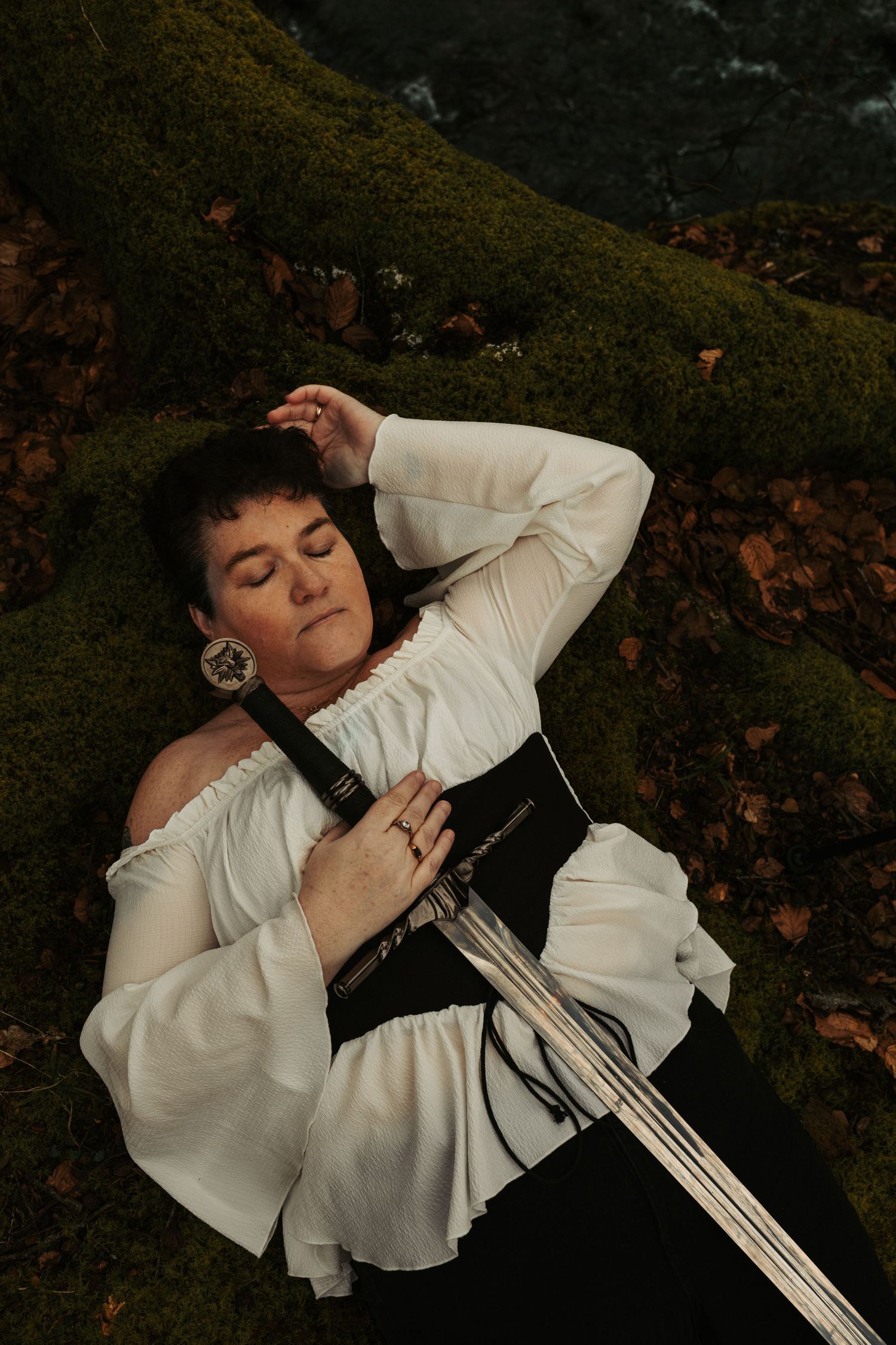 A person in medieval costume lies on mossy ground while holding a sword.