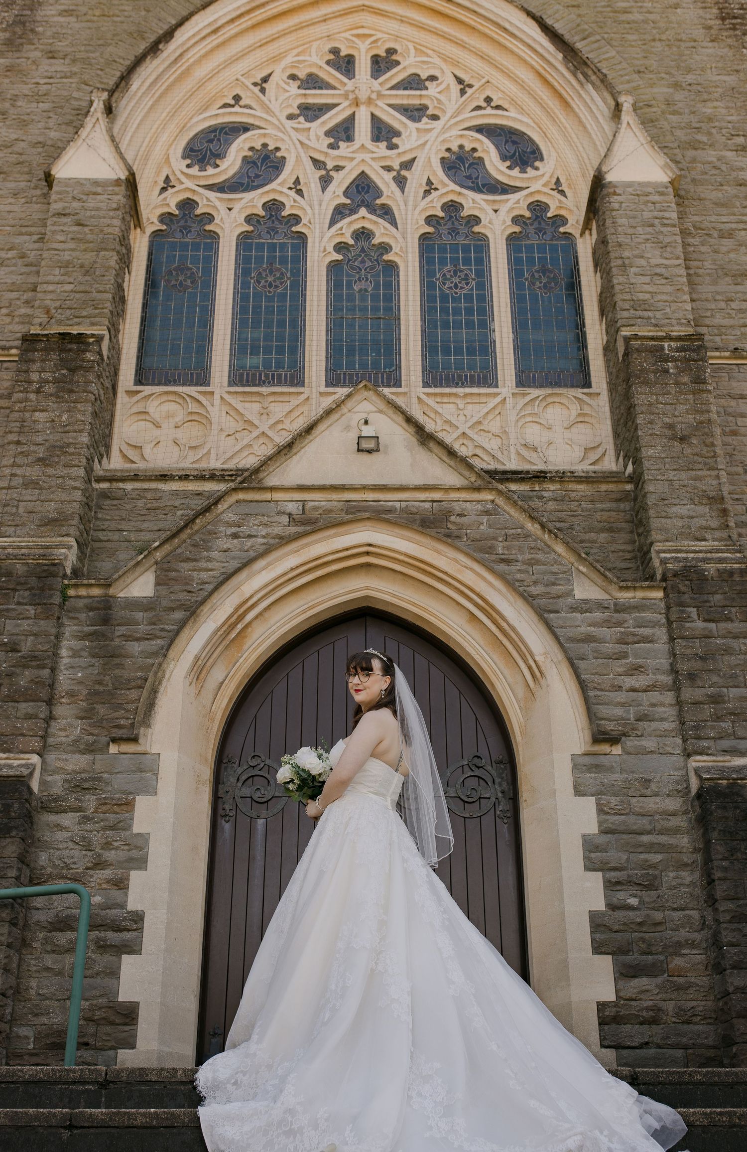 Bride in elegant ball gown poses before Gothic church arched doorway holding bouquet, veil trailing gracefully.