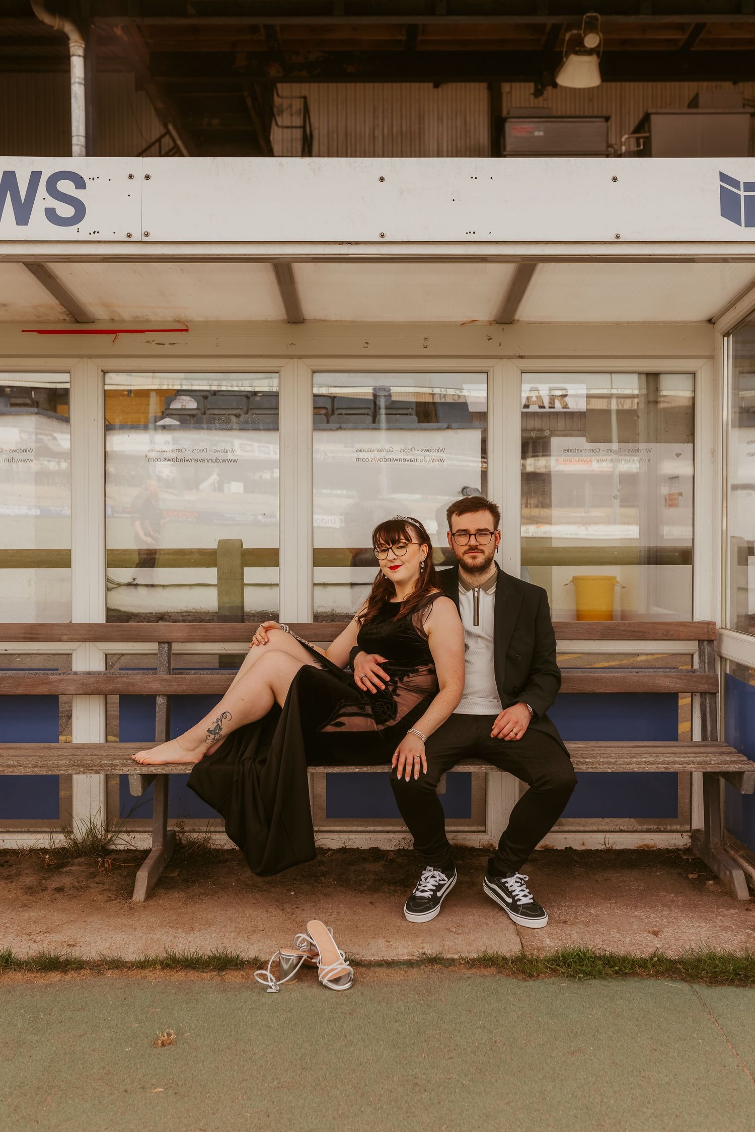 A couple in formal attire sits together on a bench at a transit shelter, the woman barefoot in a black dress.