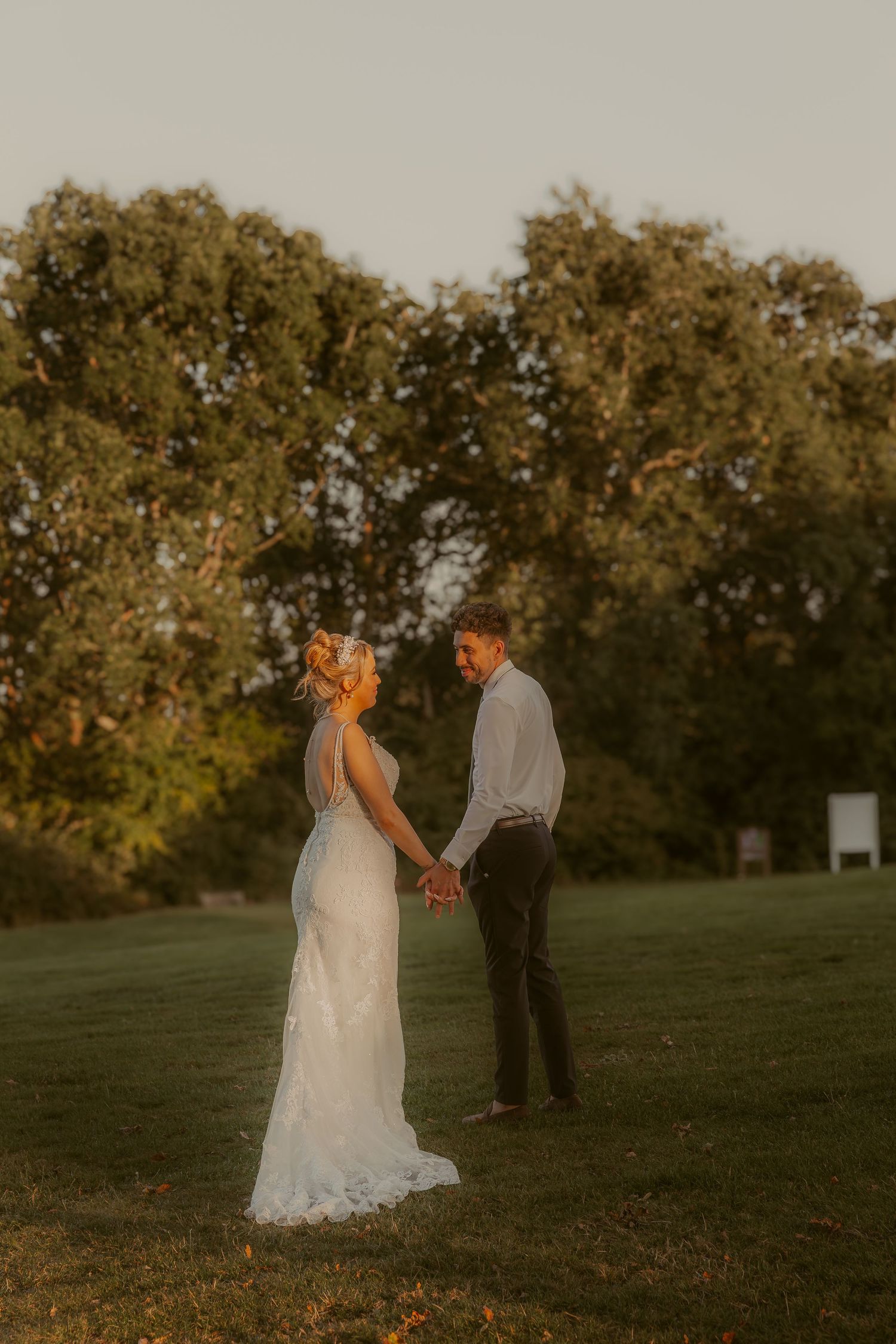 A romantic sunset wedding photo of a couple holding hands in a grassy field with trees in the background.
