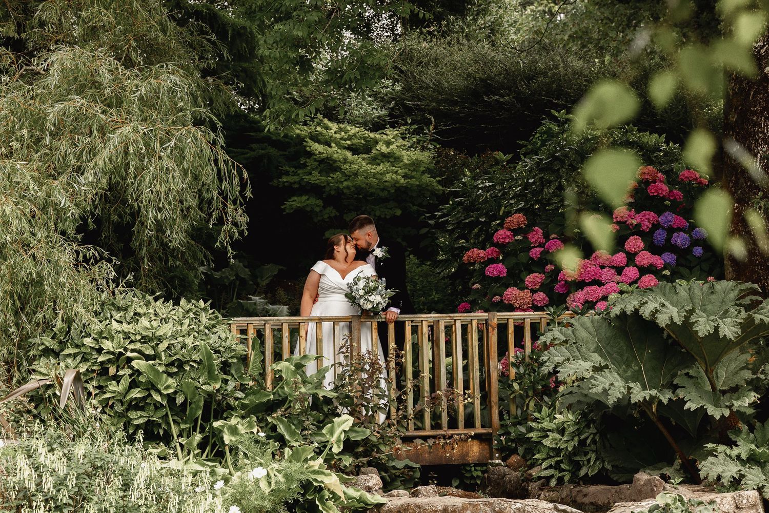 Wedding couple embracing on wooden bridge surrounded by lush garden scenery and blooming flowers.