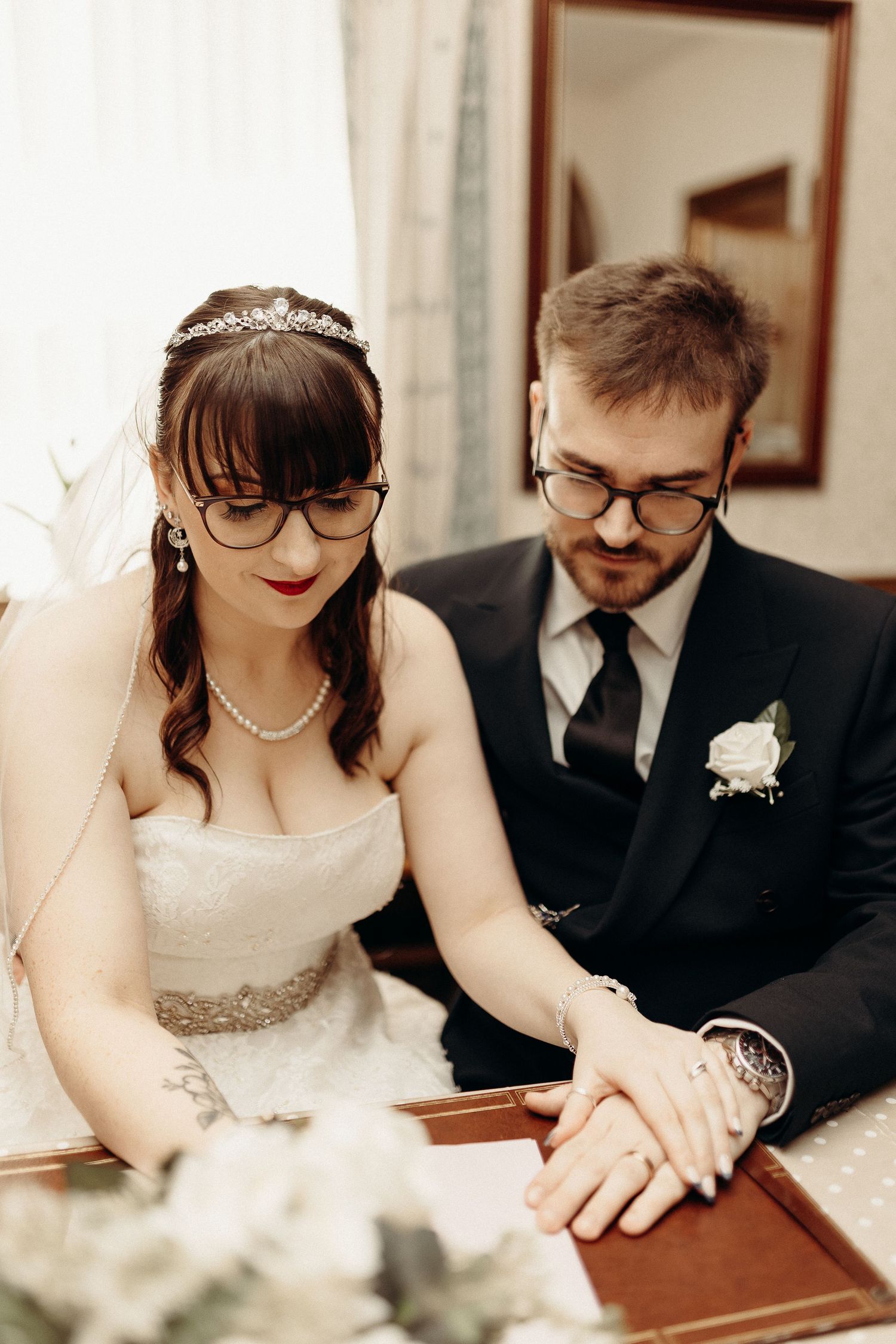 A bride and groom in glasses sign their wedding registry together, dressed in a white gown and black suit.