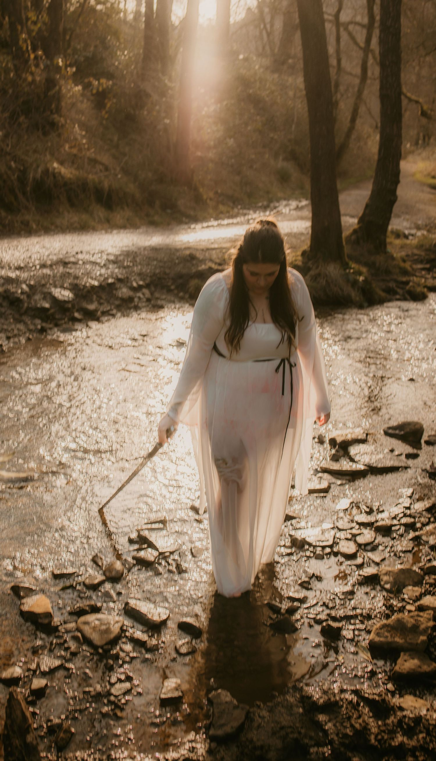 Ethereal figure in white dress walks through sunlit forest stream.