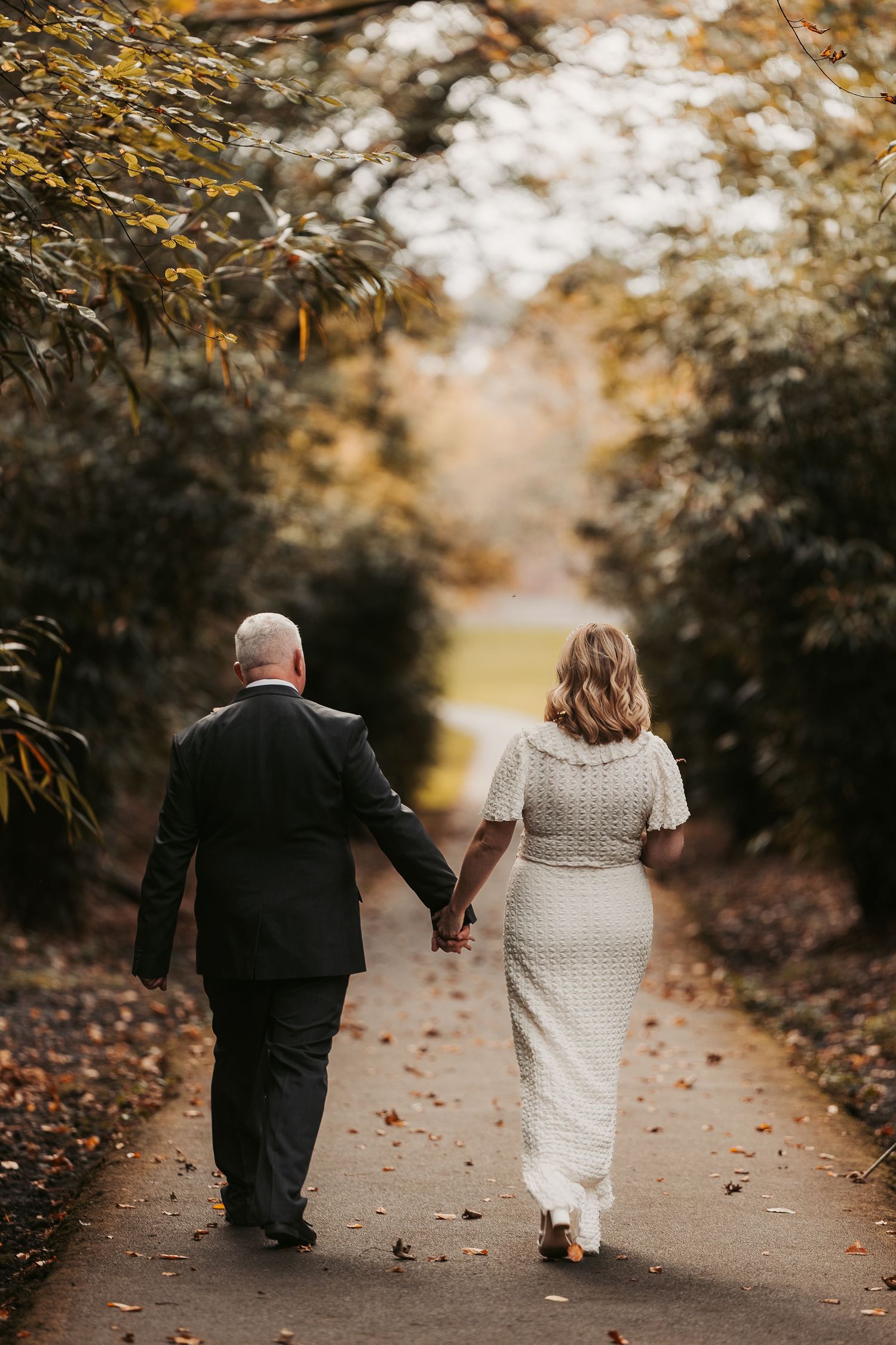 A couple walking hand in hand down a leaf-covered path in autumn.