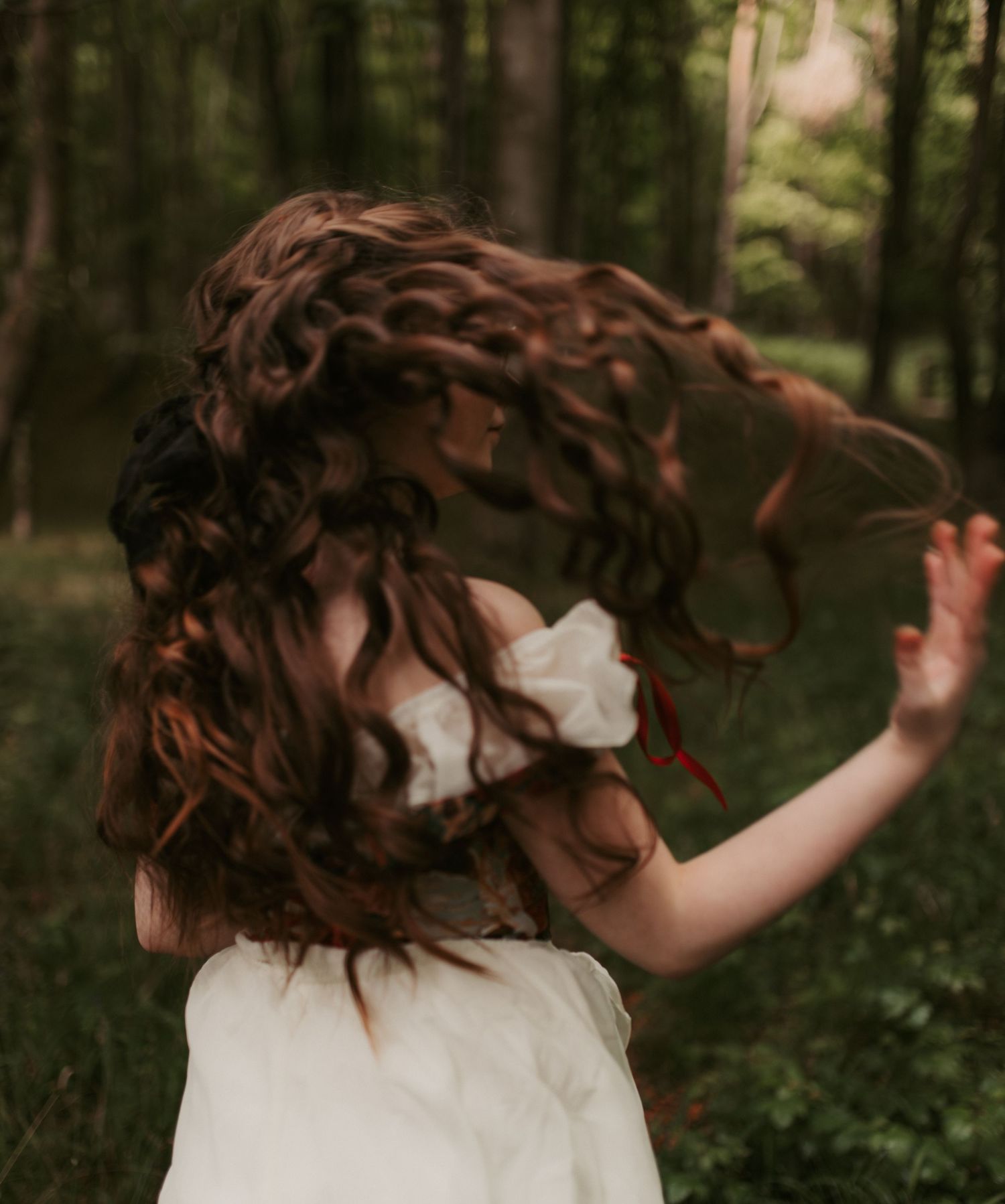 A woman in a white dress twirls in a forest, her long curly auburn hair flowing dramatically through the air.