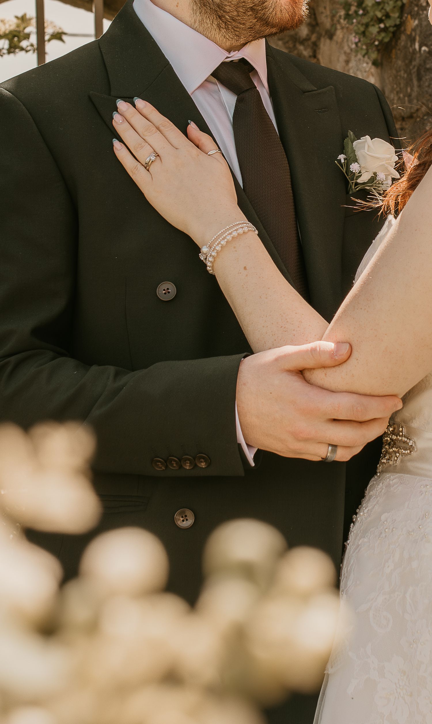 A bride and groom embrace at their wedding, showing pearl bracelet, rings, dark suit with boutonniere, and lace dress.