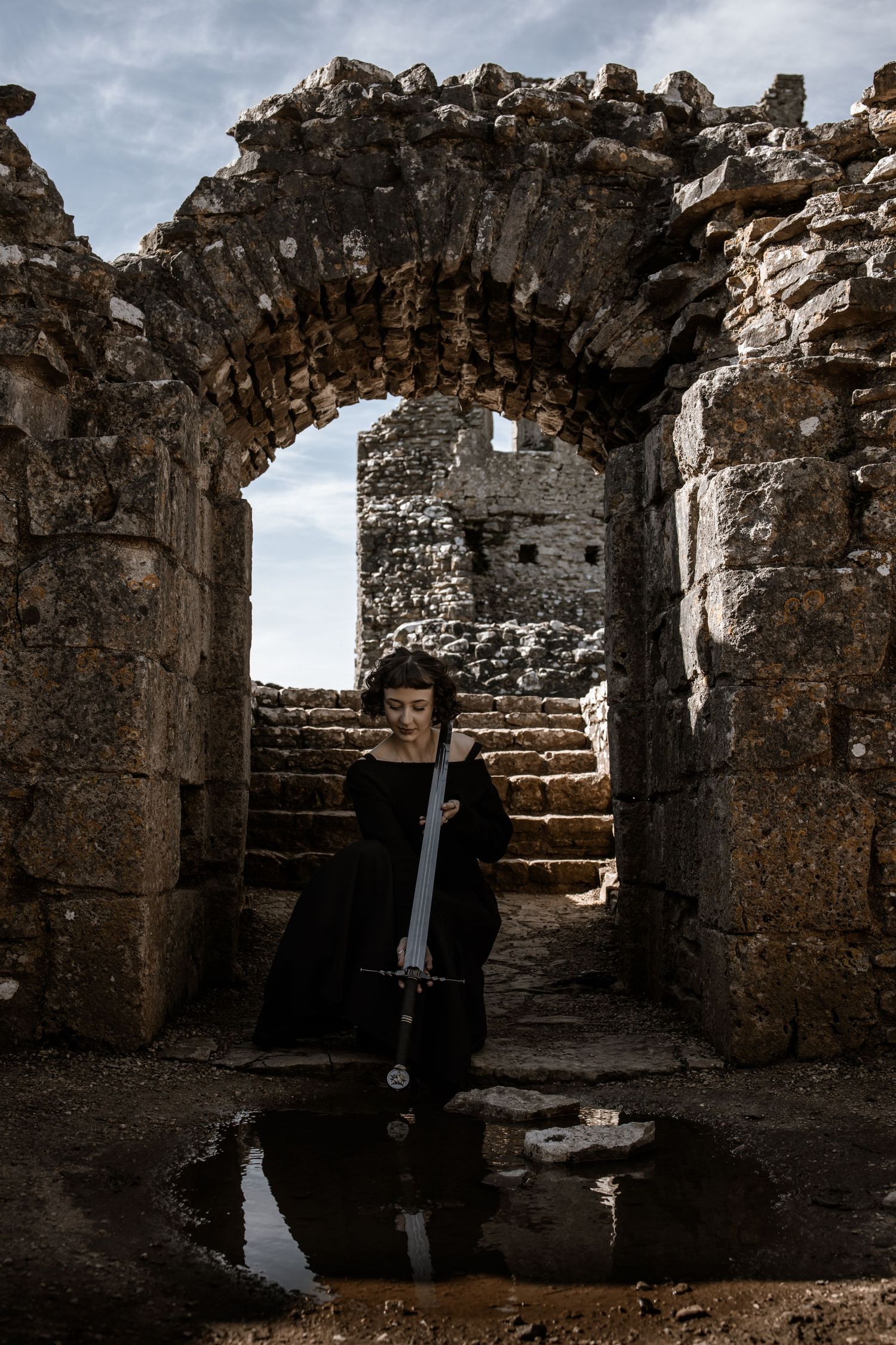 A figure in dark clothing holds a sword while standing in ancient stone ruins.