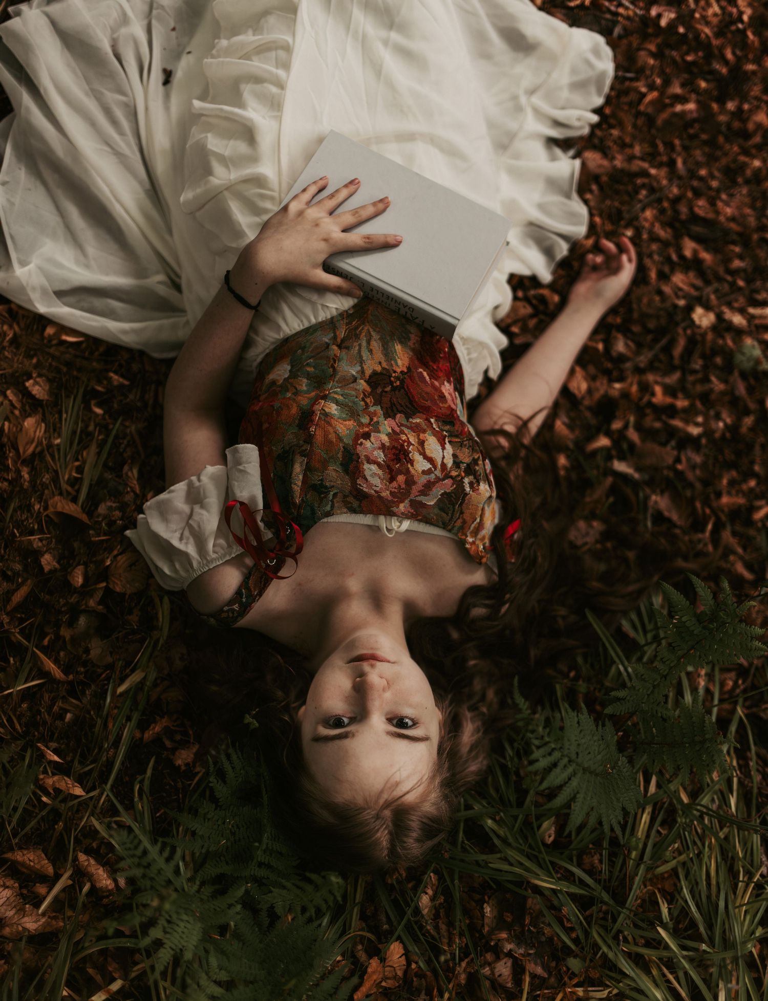 A young woman lies on autumn leaves holding a book, wearing a floral dress and white blouse, viewed from above.