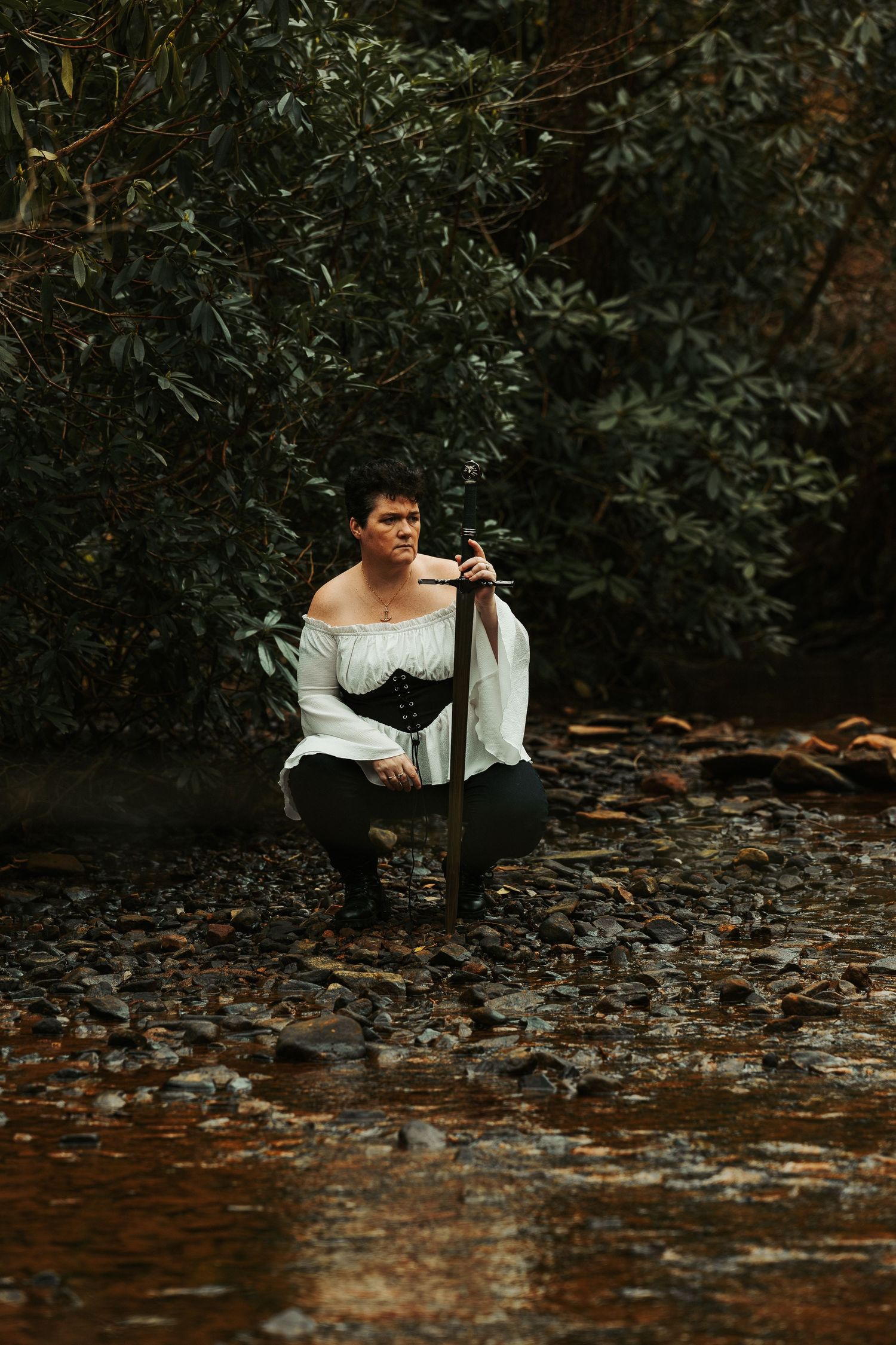 A person in renaissance clothing crouches by a stream in a moody forest setting.