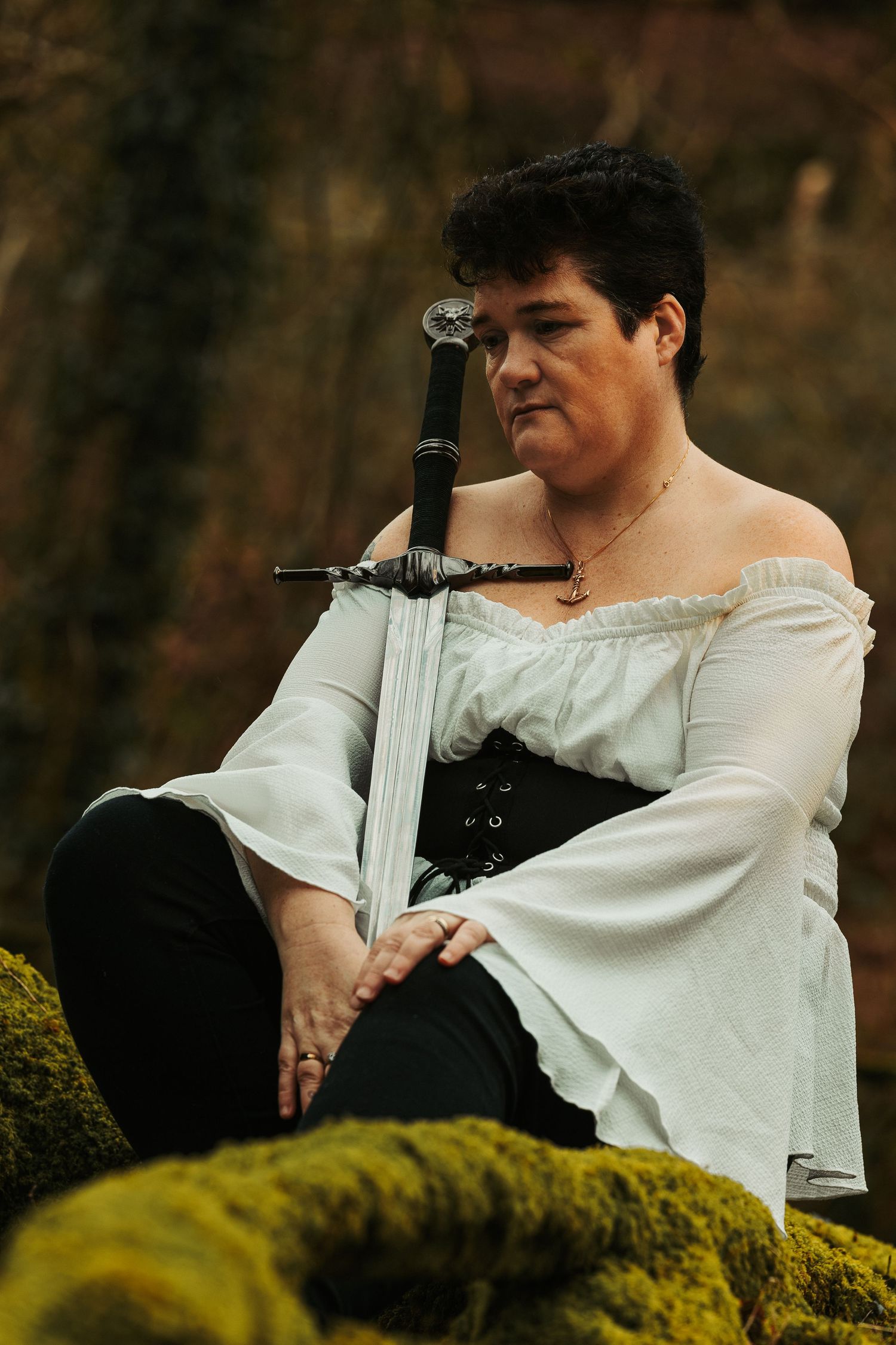 A figure in white period clothing poses with sword against mossy natural backdrop.
