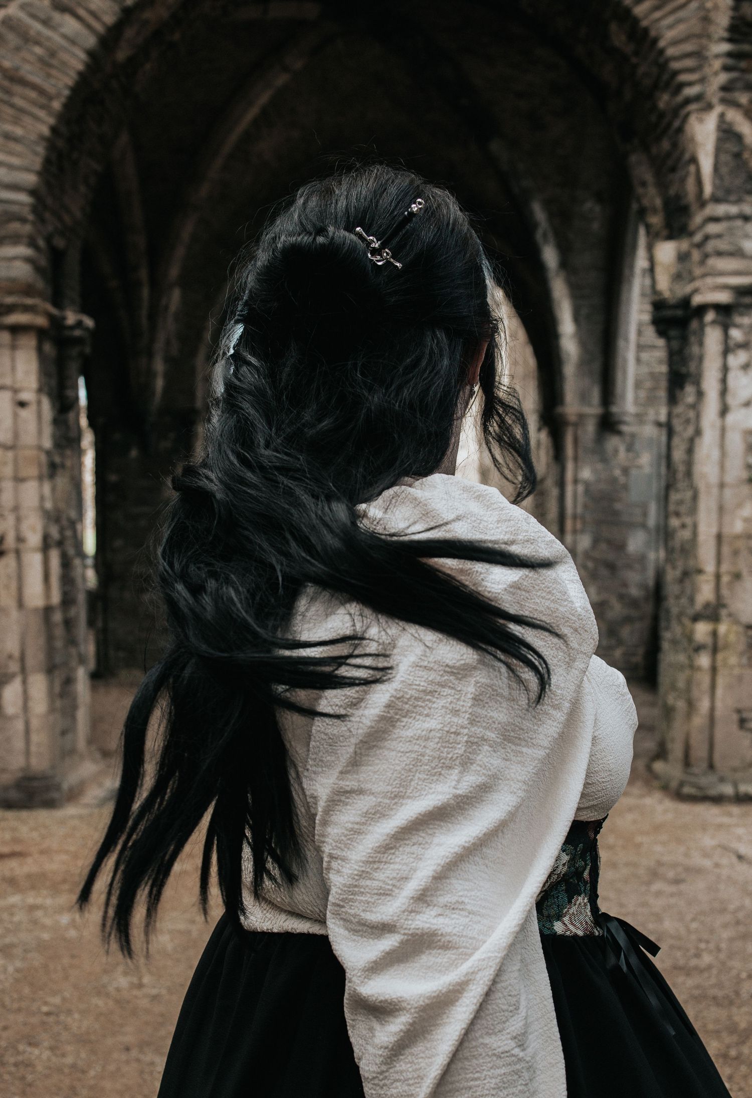 A person with long dark hair wearing medieval-style clothing stands beneath stone archways.