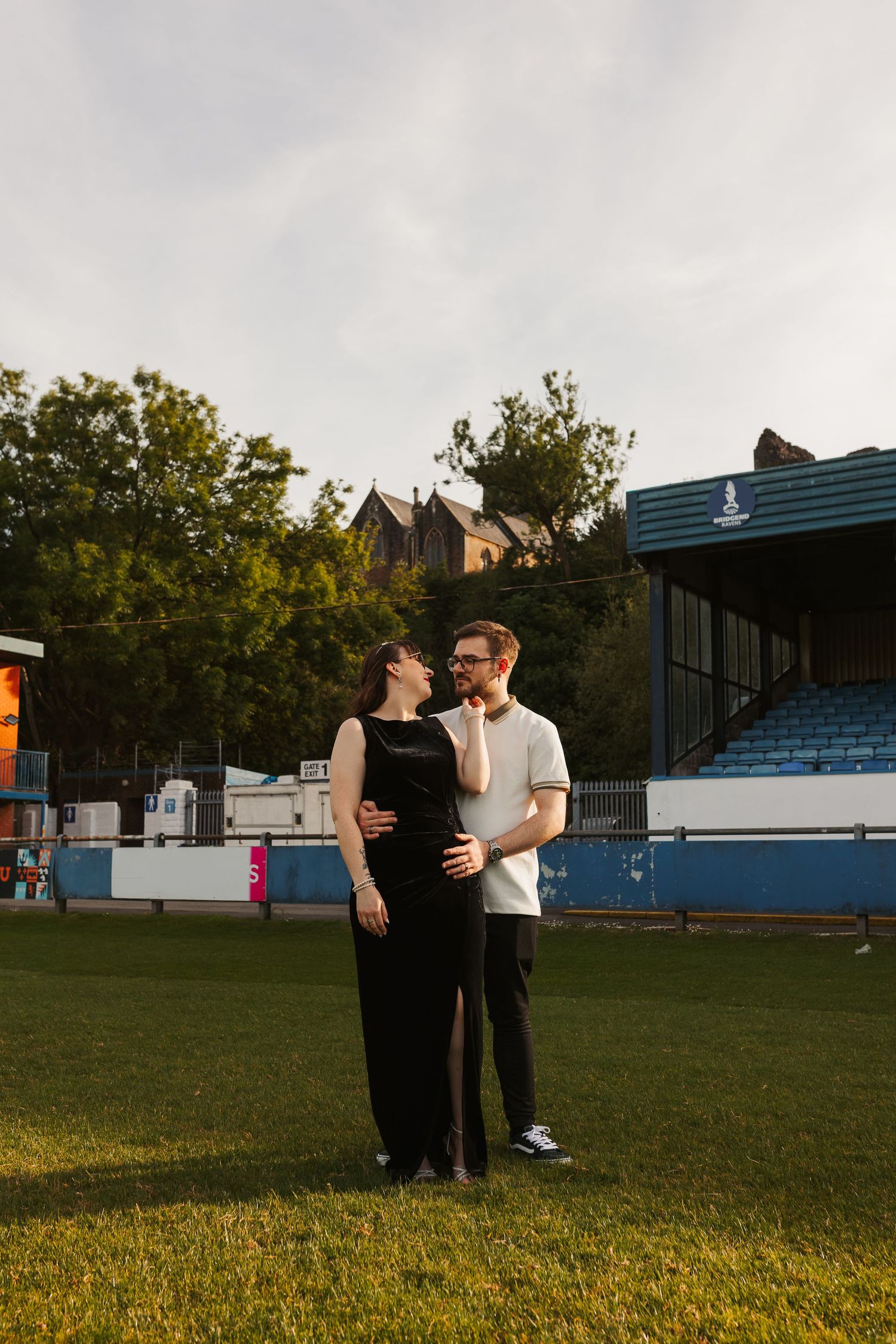Couple embracing on a grass field at a stadium during golden hour, man touching pregnant womans belly.