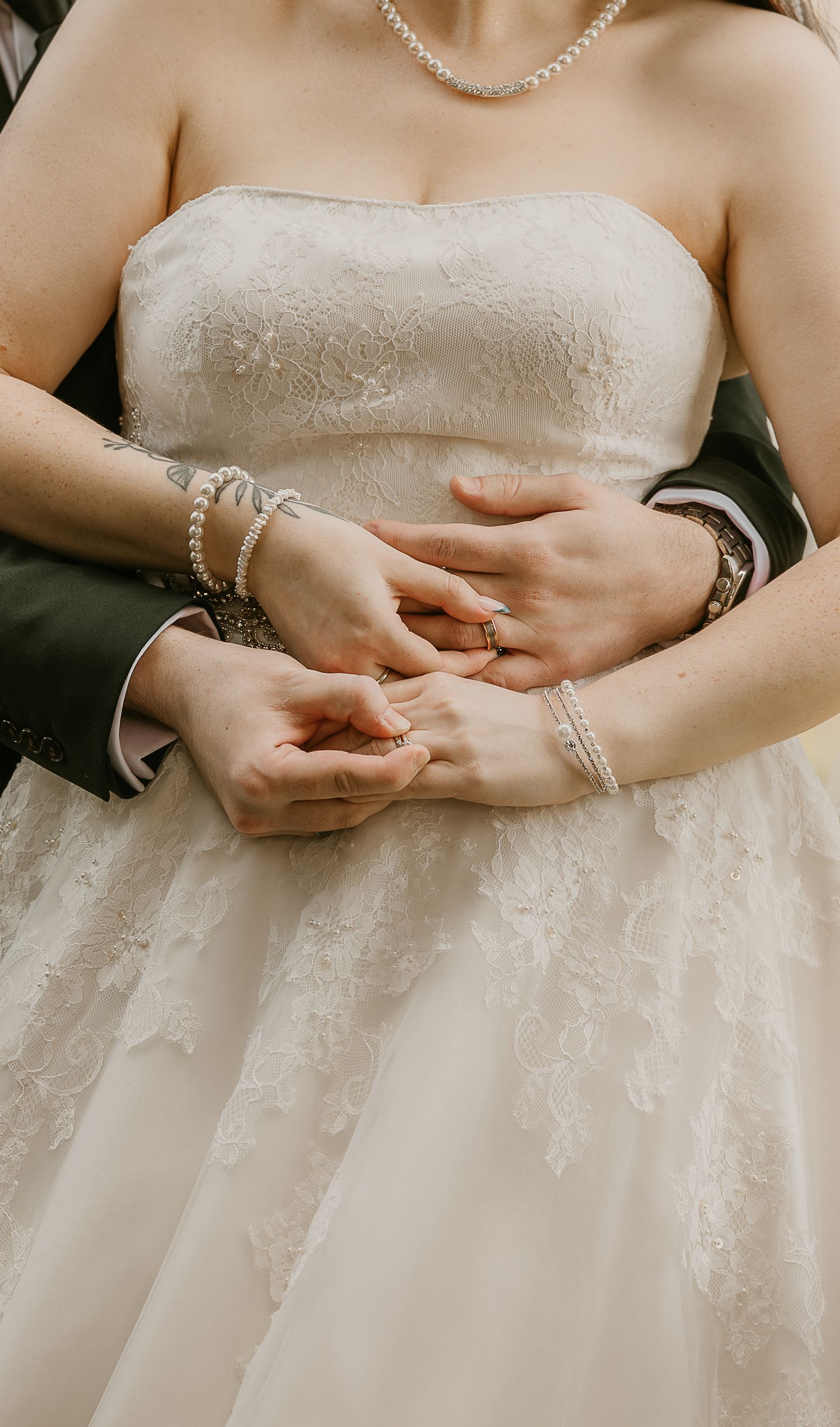 Bride and groom exchanging wedding rings, bride wearing strapless lace gown and pearl necklace, groom in dark suit.