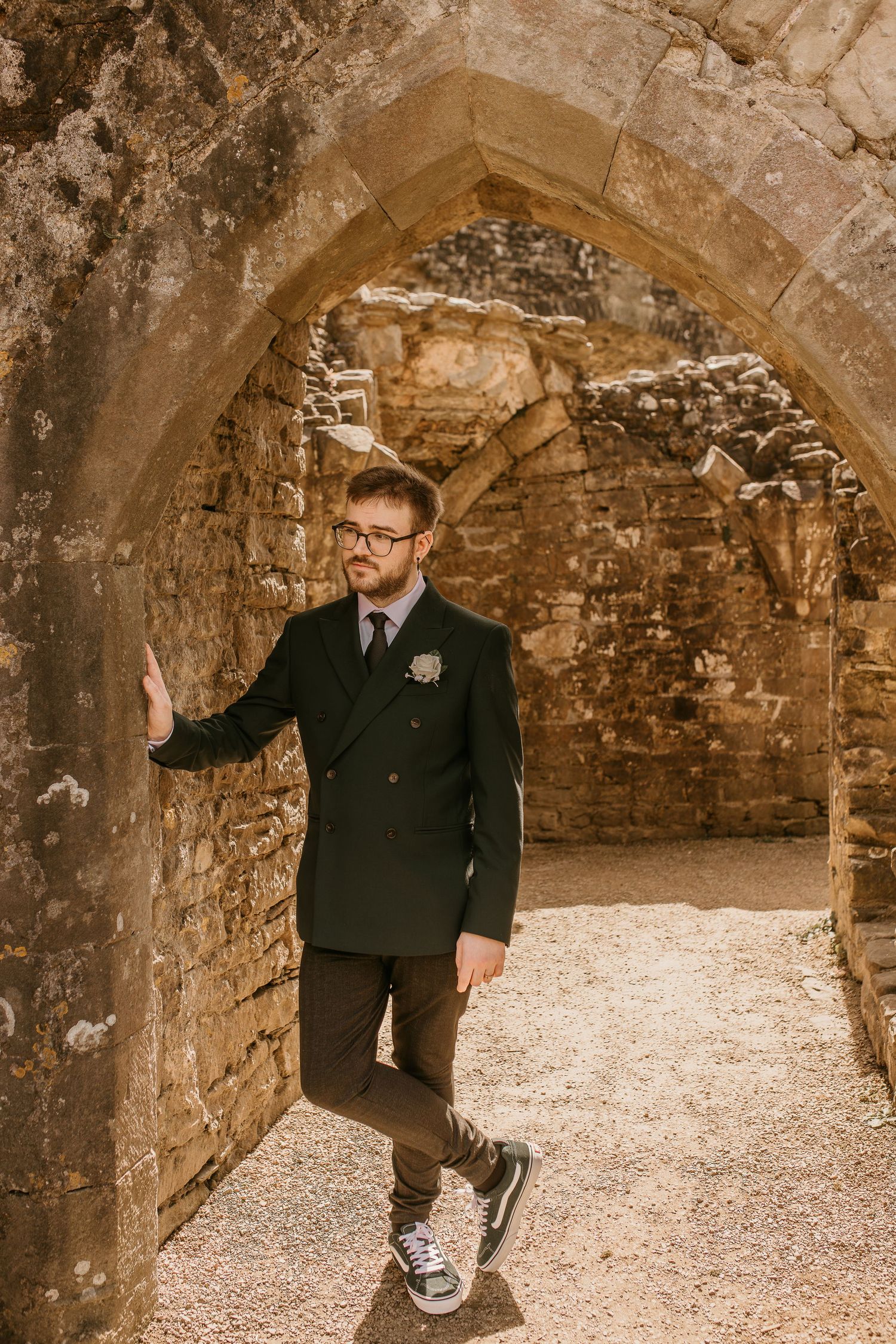 Groom in black suit stands under stone arch at historic ruins.
