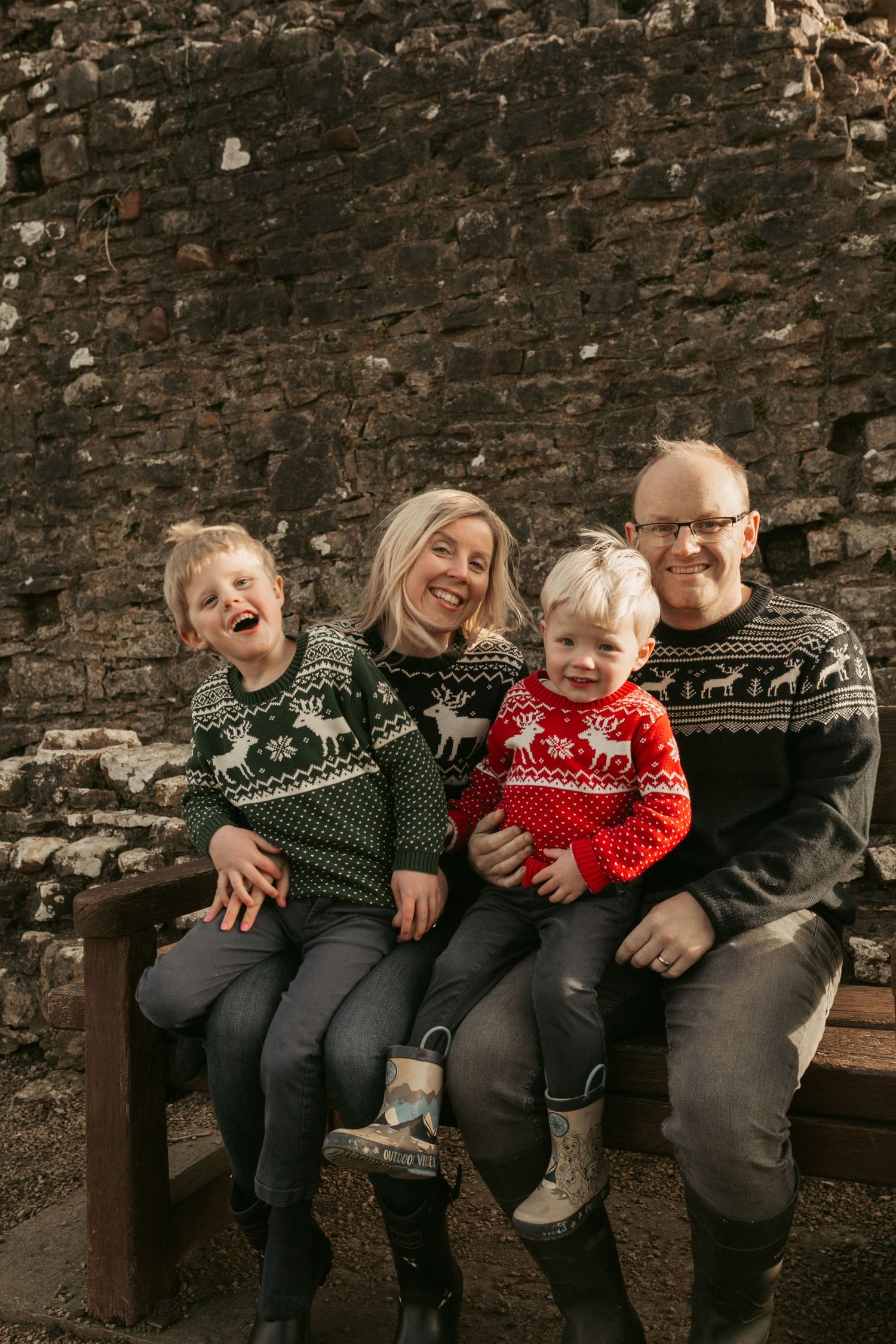 A happy family wearing matching winter sweaters sitting together on a bench against a brick wall.