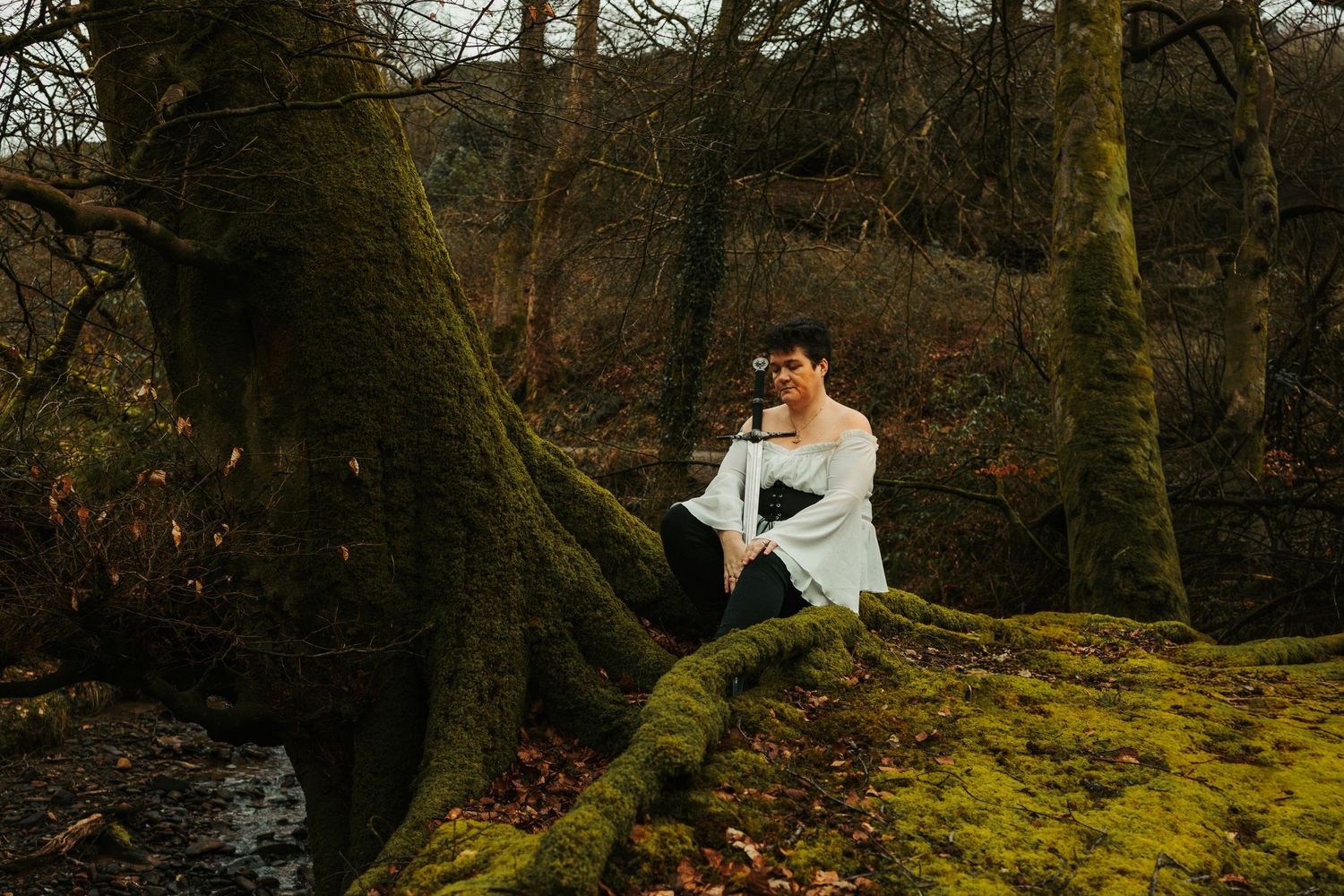 A person in white blouse poses dramatically on moss-covered ground in autumn forest.