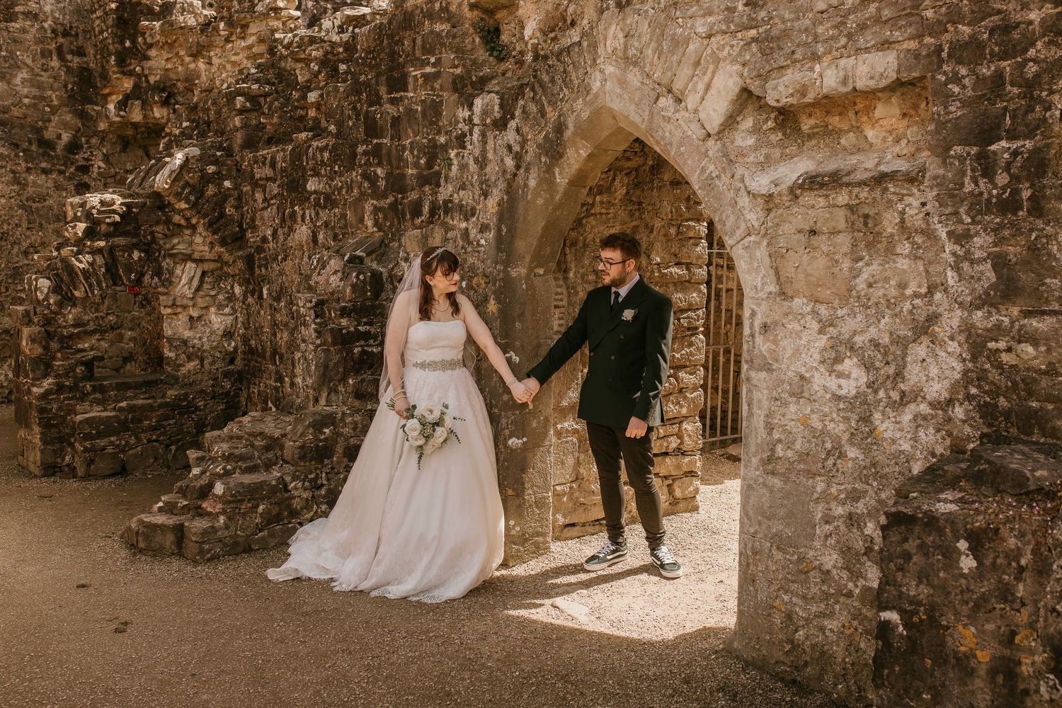 Bride and groom hold hands under stone archway at historic ruins wedding venue.