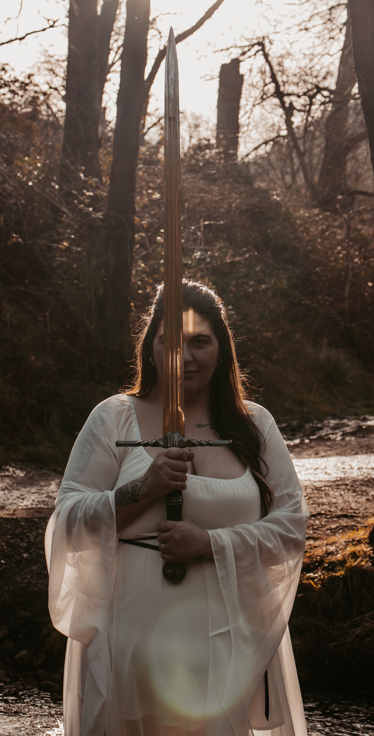 A figure in a flowing white dress holds a sword in a misty forest setting at sunset.