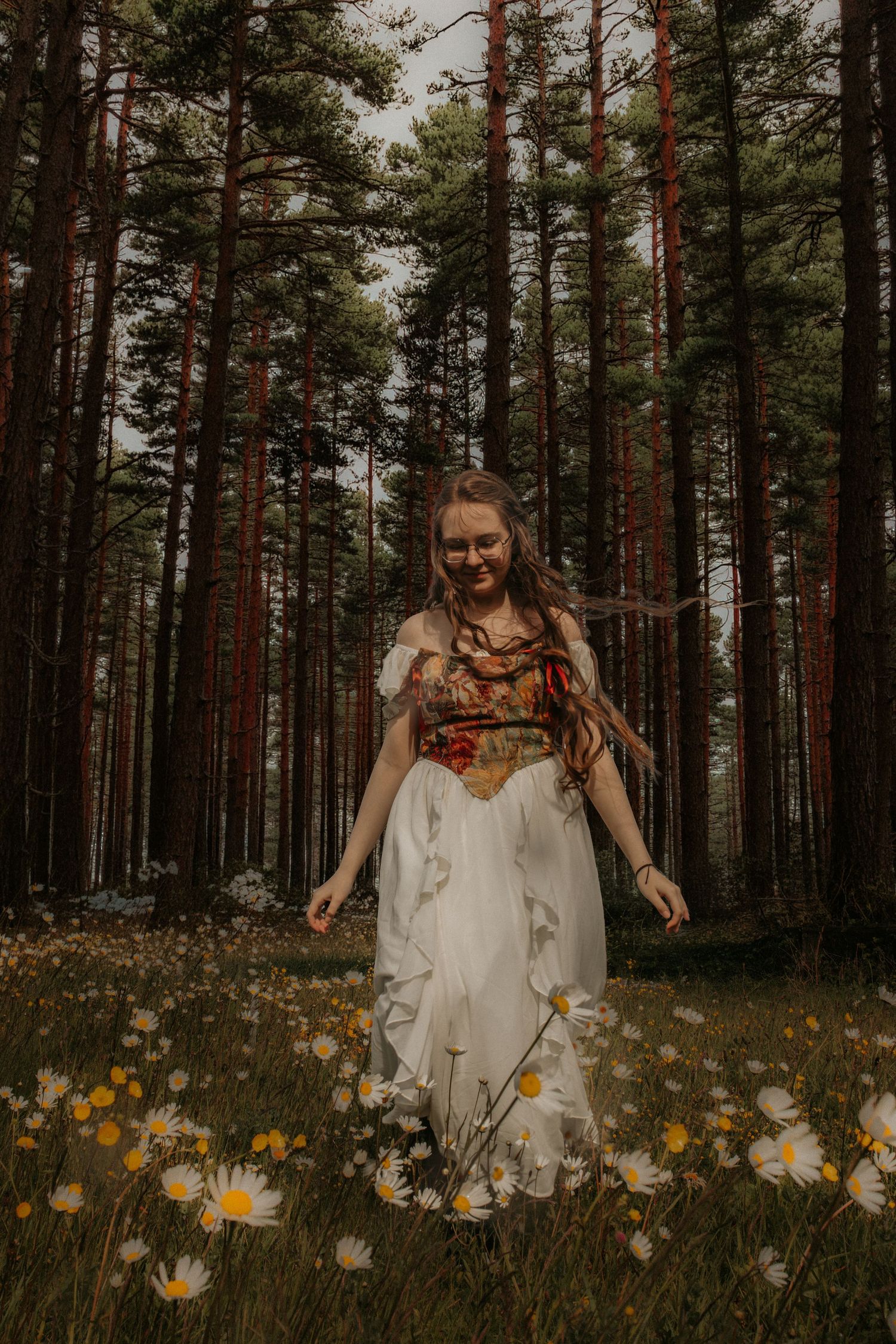 Young woman in floral bodice and white skirt walks through wildflower meadow surrounded by tall pine forest.