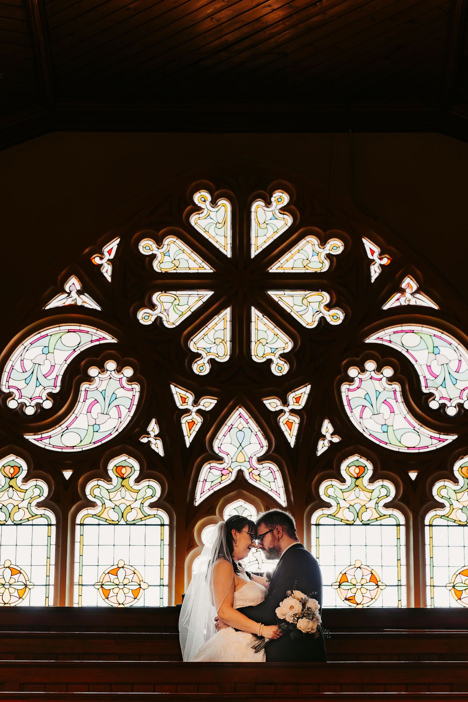Bride and groom sharing an intimate moment in front of a stunning stained glass rose window in a church.